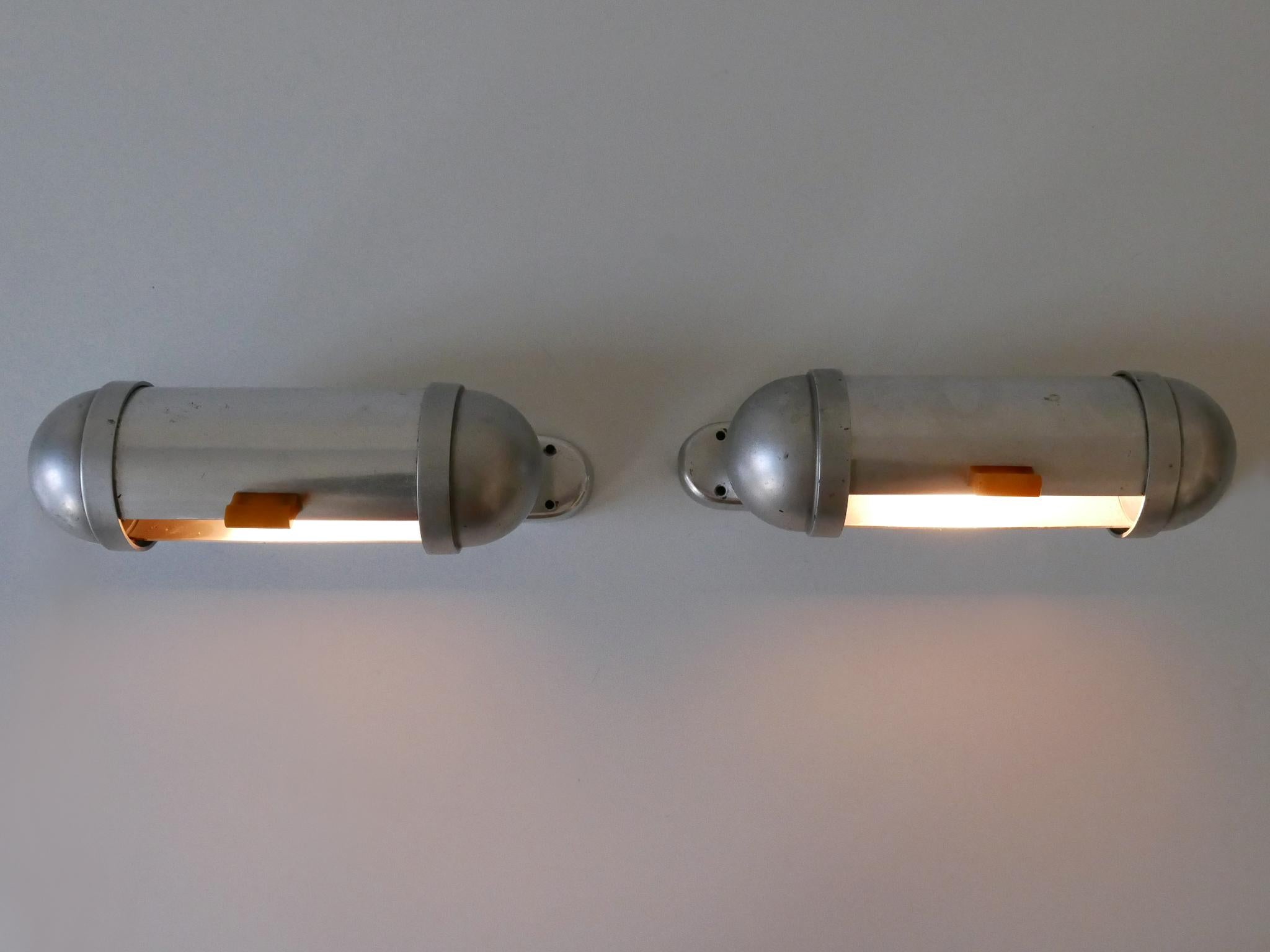 Set di due eleganti e minimali appliques dal design filante per armadietti sottomarini / u-boot di König, Germania, anni '40. Inciso sul retro.

Realizzato in alluminio e bachelite, ogni apparecchio è dotato di 1 portalampada a vite E27 / E126 di