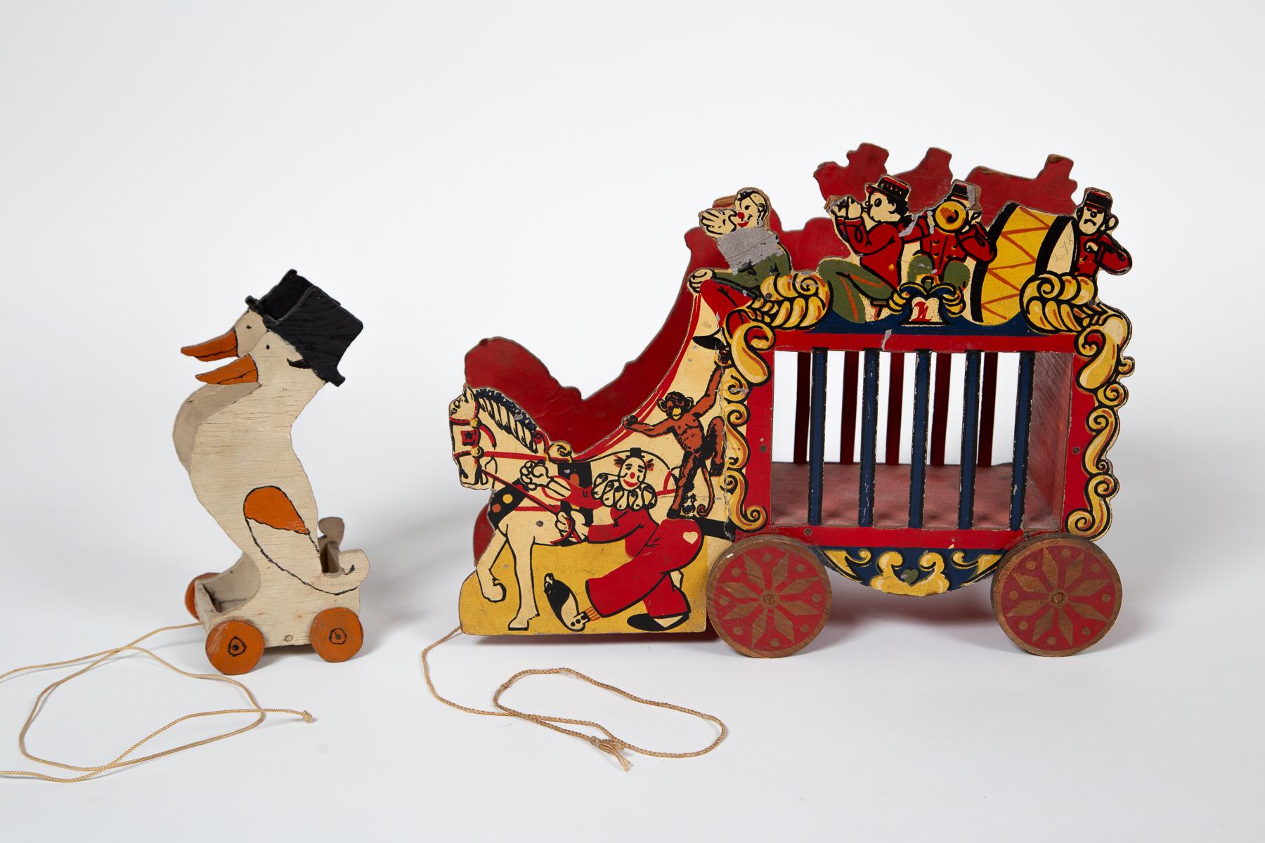 Satz von zwei Holzspielzeugen, 20. Jahrhundert. Charmanter Volkskunst-Zirkuswagen und Entenpaar. Zirkuswagen mit aufgelegter Lithografie der auftretenden Figuren. Die Enten mit Zylinder sind handbemalt. Beide Stücke haben funktionierende Holzräder