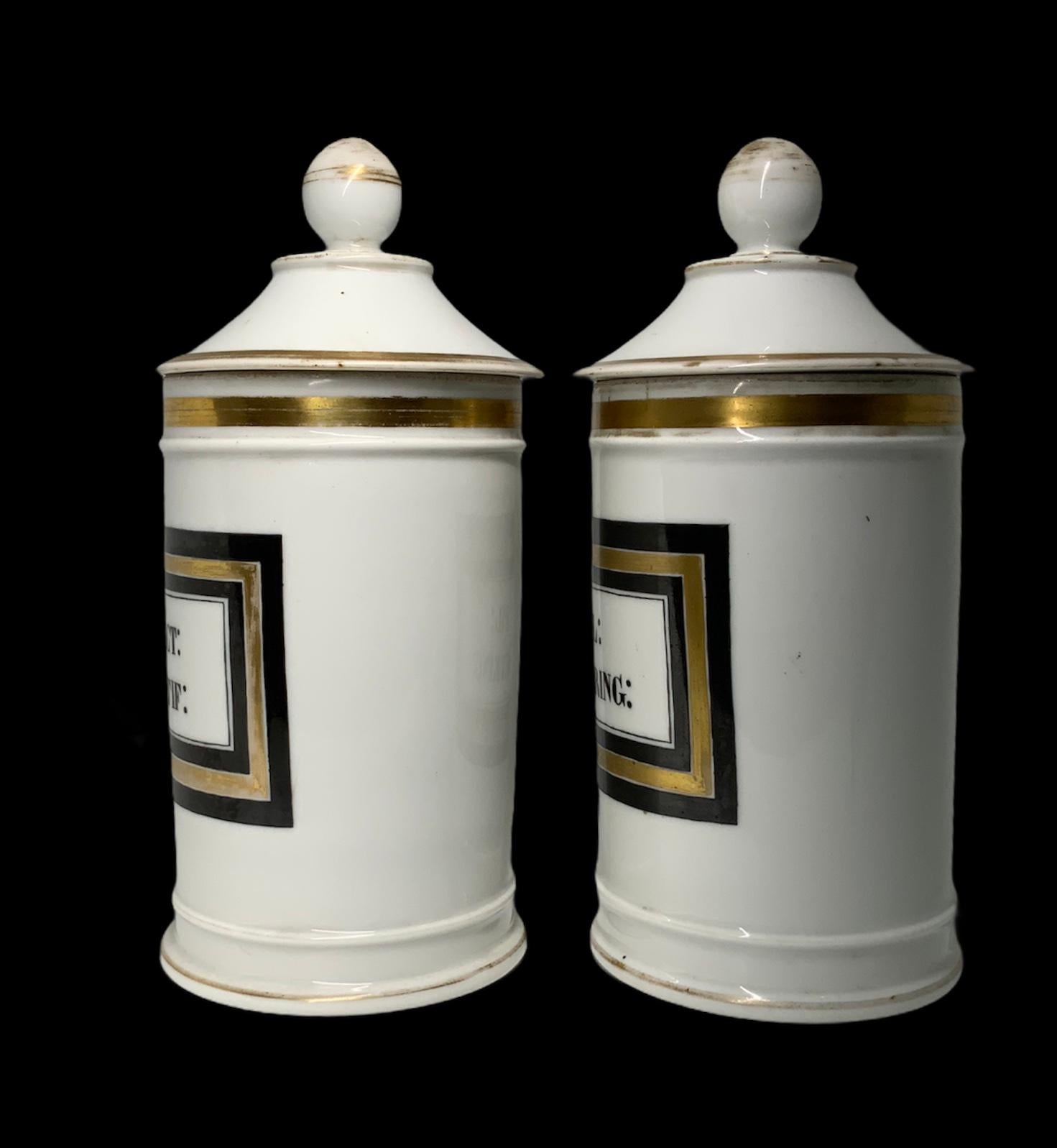 This is a pair of porcelain apothecary cylindrical jars with lids. The color of their background is white and they are decorated with gold bands in their upper and lower border. There are a rectangular plaque with hand painted black and gold borders