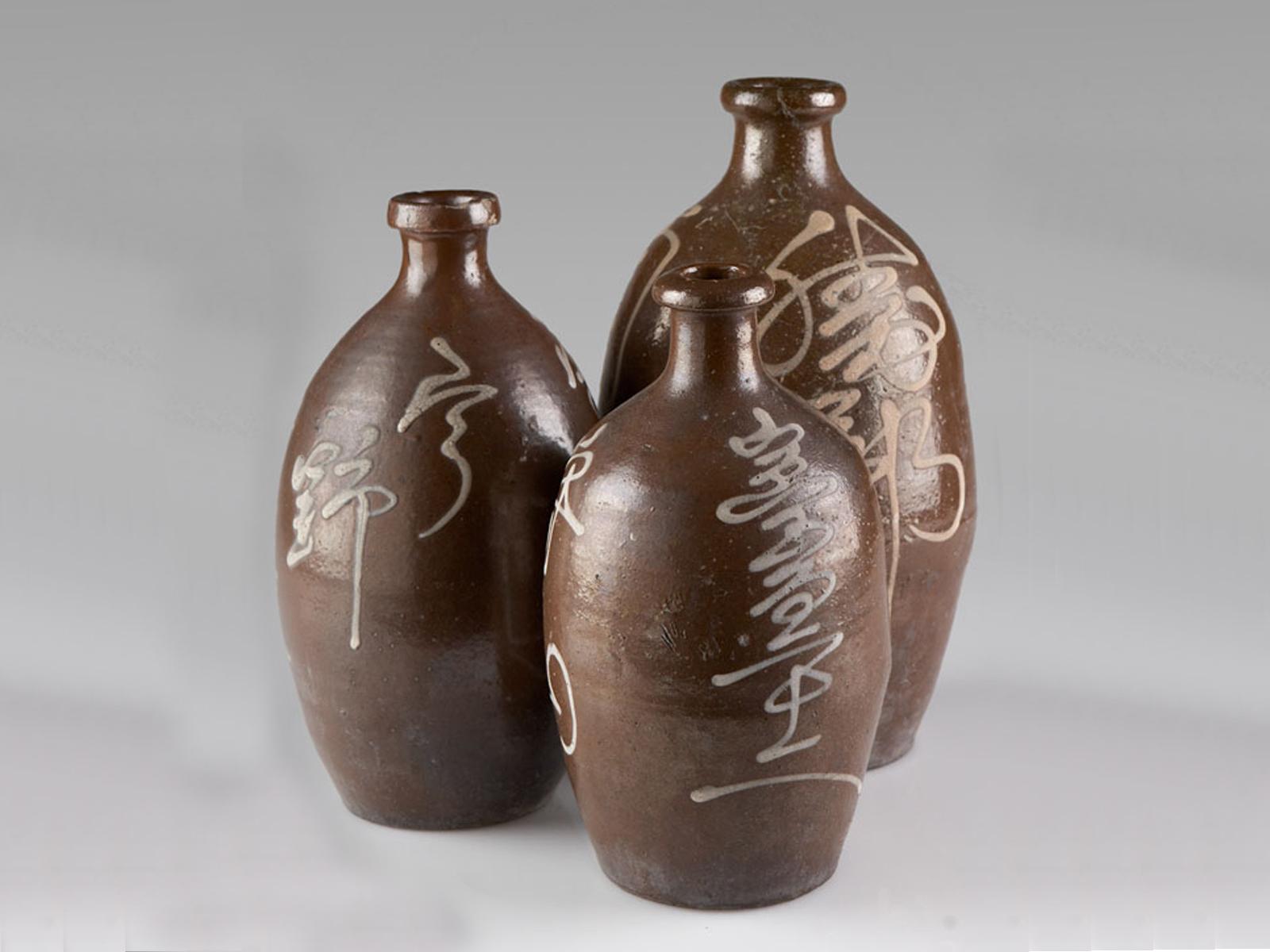 A trio of antique Japanese handmade earthenware saki sake bottles from Japan, circa 1900. Each jar has a different signature in script across the front that is the mark of the master saki maker responsible for the contents. The thick rim was sealed