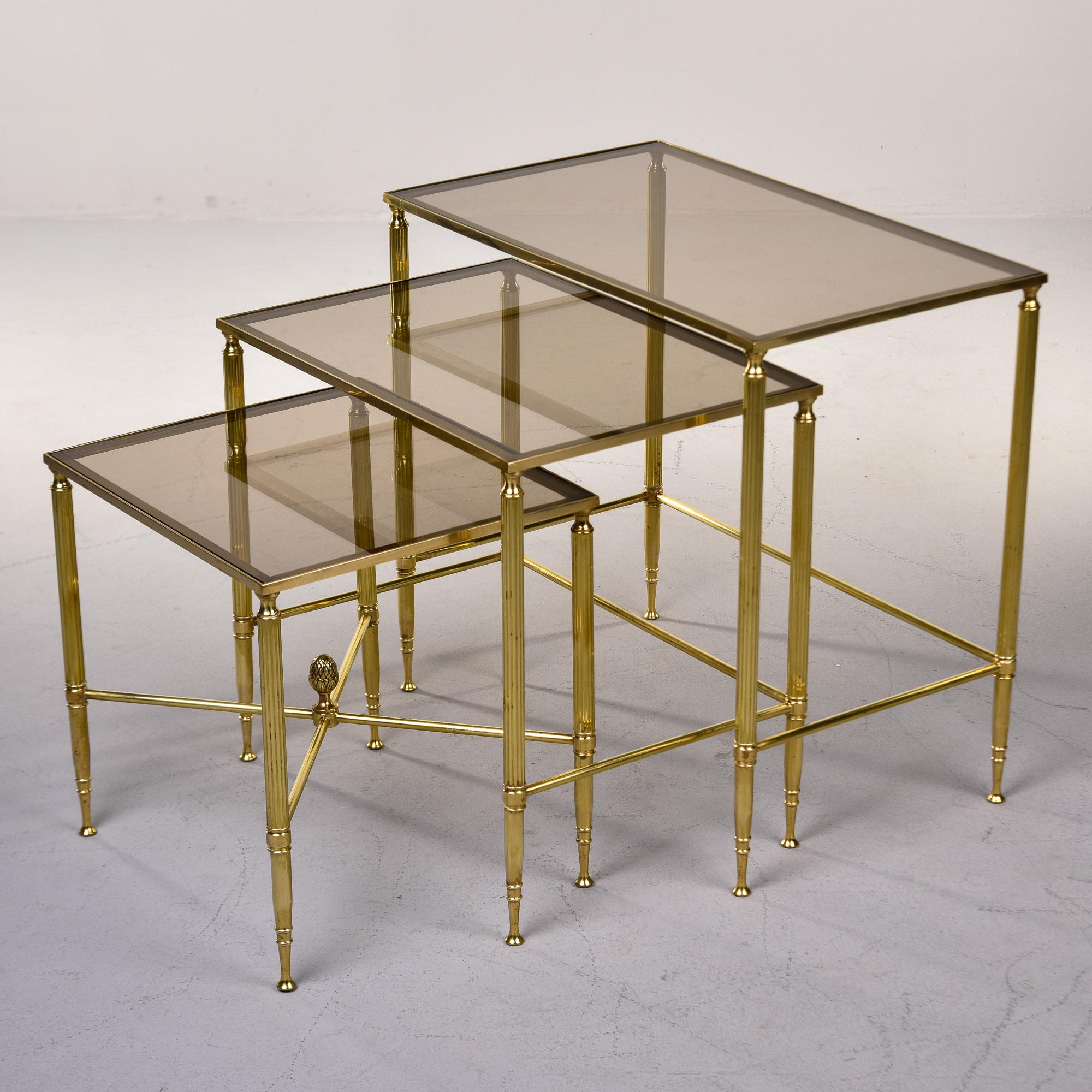 Found in the US, this set of three brass and glass neoclassical nesting or stacking tables dates from the 1970s. Each table features a brass frame with reeded, tapered legs and a table top insert of taupe-tinted glass. Smallest table of three has