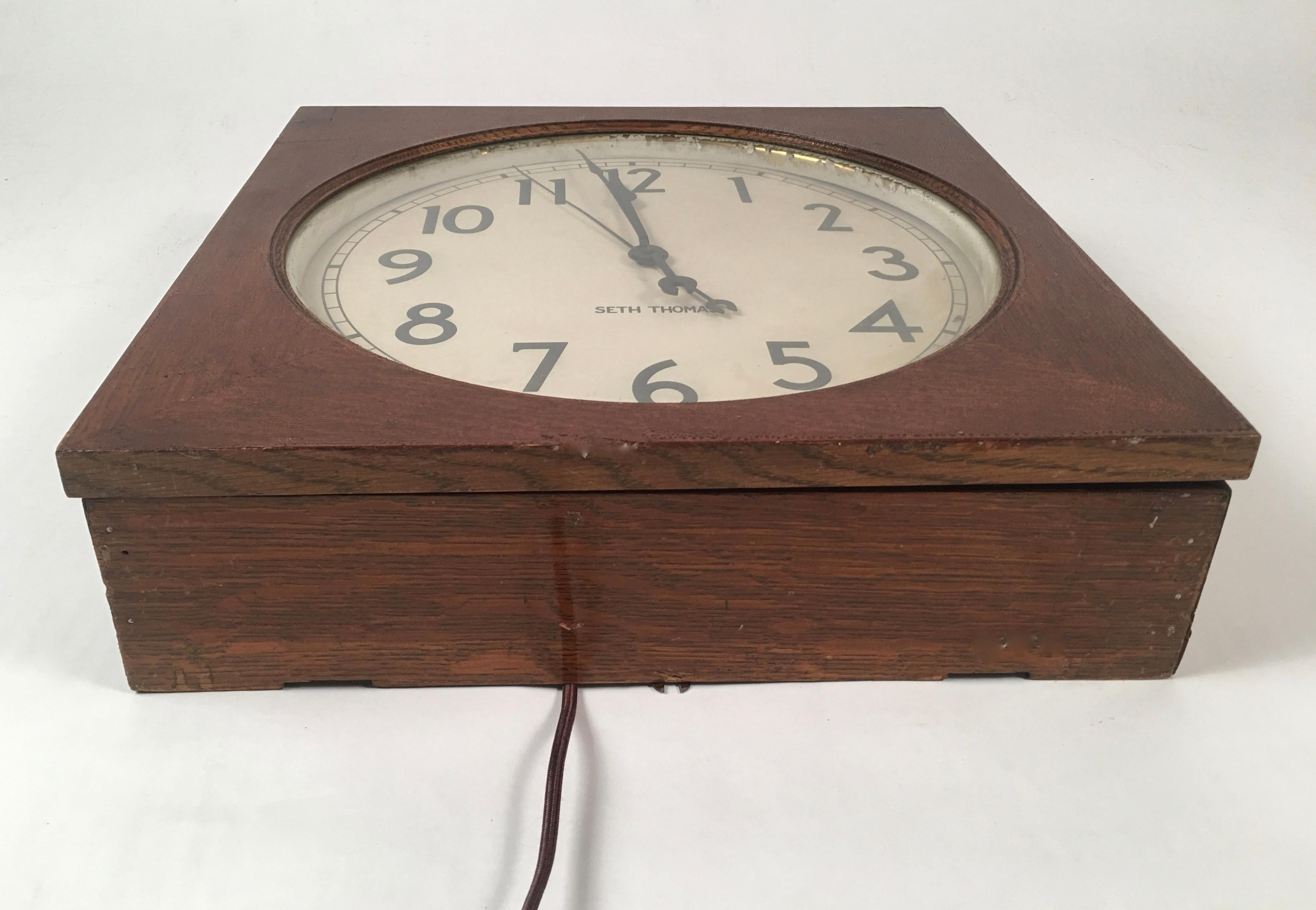 A Seth Thomas electric wall clock, the square case with its original varnished surface, now wonderfully leathery, with a hinged cover with a round glass window which opens up to the white painted metal dial with black numerals. Metal plaque inside