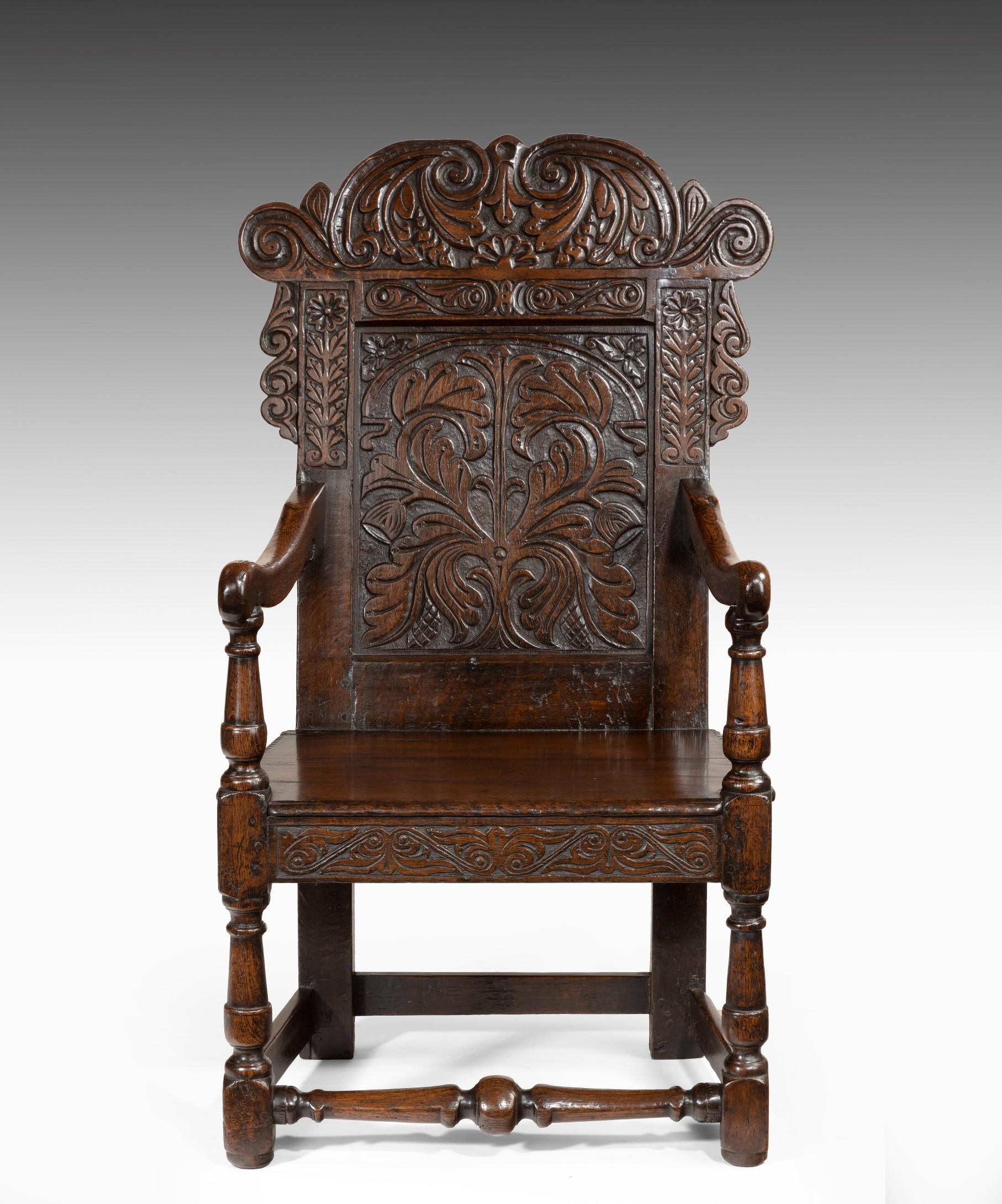 A profusely carved late 17th Century oak wainscot armchair. The armchair's scrolling foliate leaf cresting rail above a central panel carved with flowing foliage and flanked by uprights carved with palmettes; having downswept arms and a boarded seat
