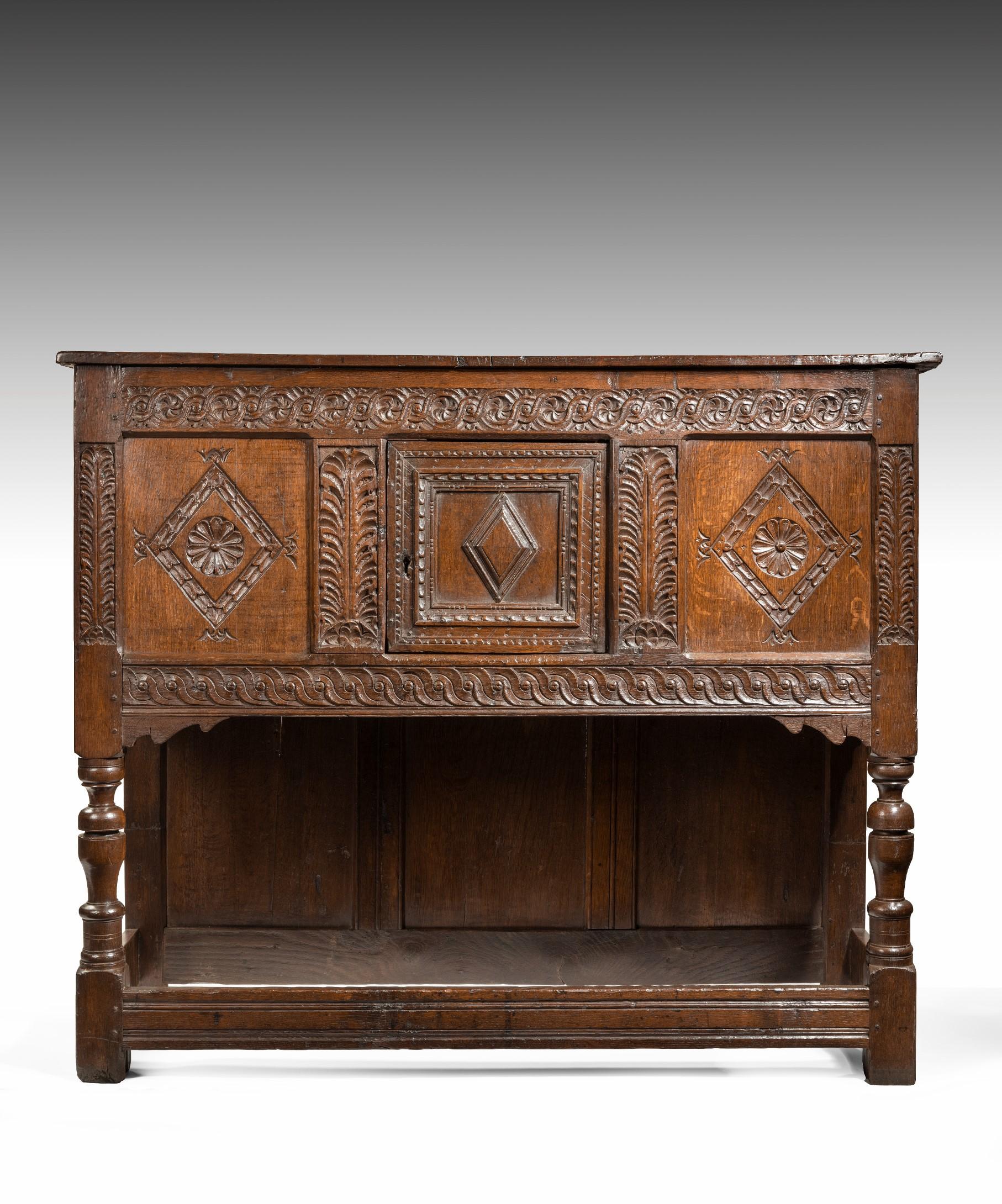 A handsome 17th century Charles II oak livery cupboard with wonderfully bold carving; the cupboard's boarded top above a three panelled front with a central door and raised on cup and cover turned legs united by moulded stretchers. The livery