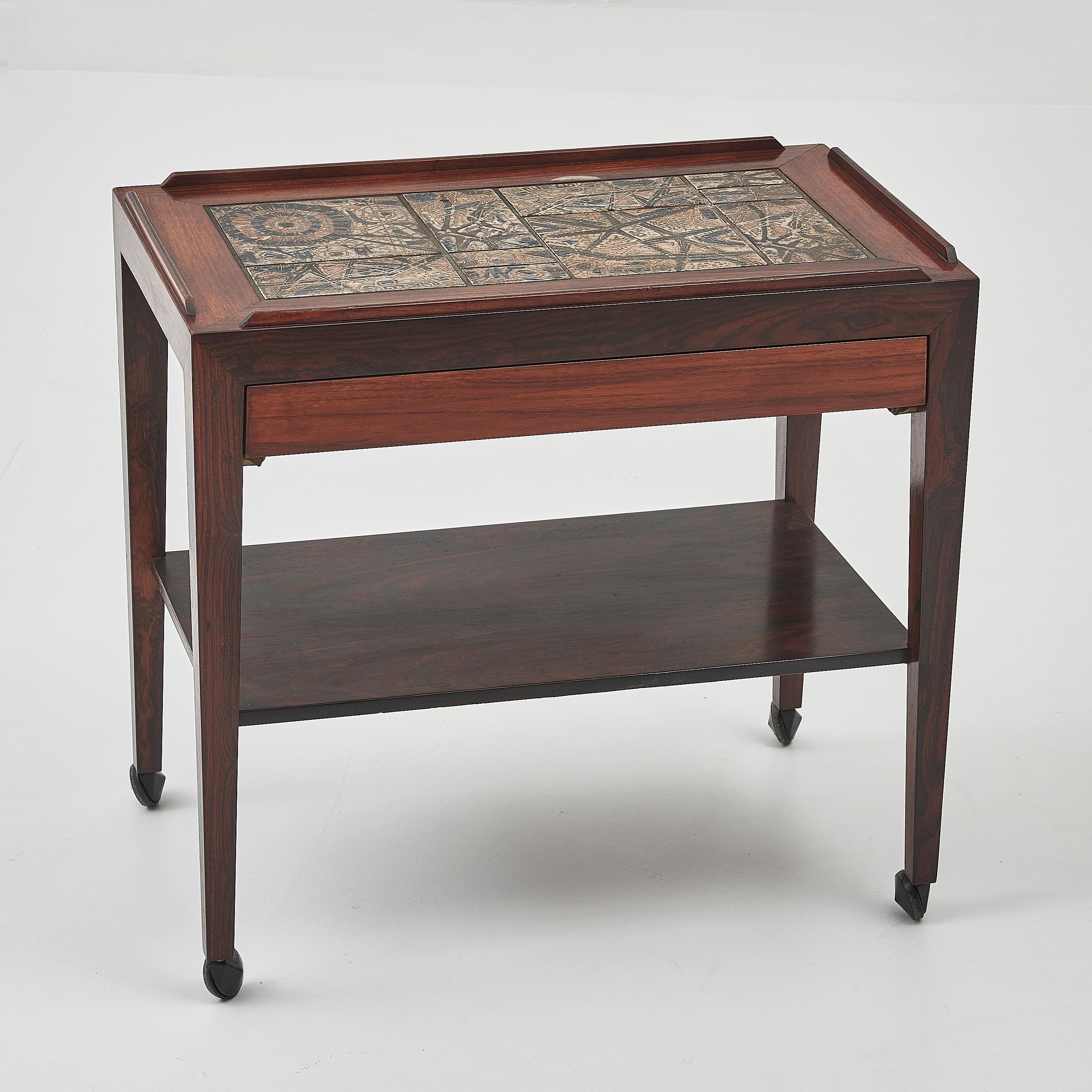 The Severin Hansen rolling rosewood side table is a masterful blend of Danish design and artisanal craftsmanship. Featuring a sleek rosewood frame, this table is elevated by a stunning ceramic tiled top, hand-painted and glazed by artists in