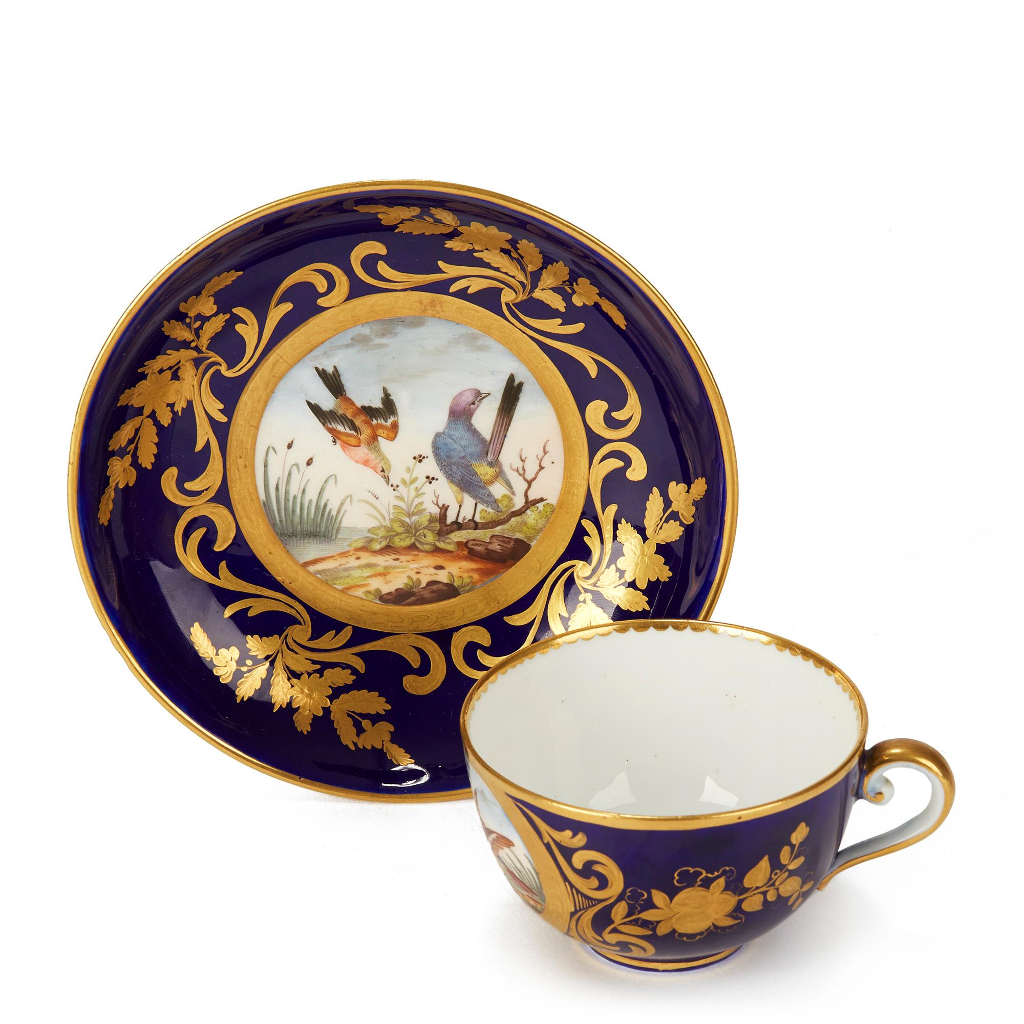 An exceptional and rare Sèvres Porcelain cabinet teacup and saucer each hand painted with birds within a landscape in coloured enamels and set within a cobalt blue surround exquisitely highlighted with gilding patterns including scrolling leaf