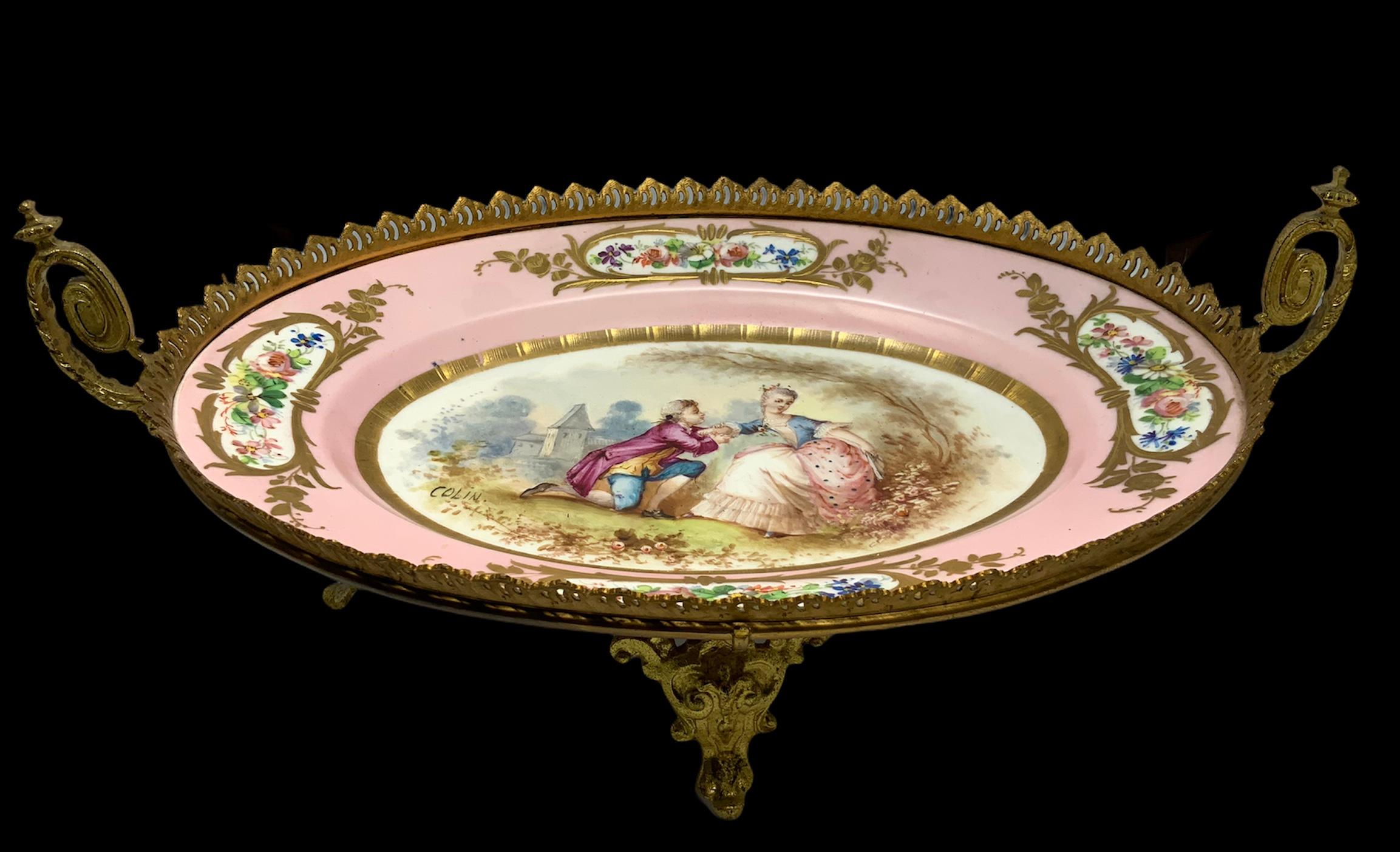 An 1846 Sevres Porcelain hand painted with a romantic couple scene in a pastoral setting and signed by Colin in the center of a pink dish. Adorned with flowers and gold acanthus leaves and mounted in a bronzed pierced ruffle border. Gilt bronze coil