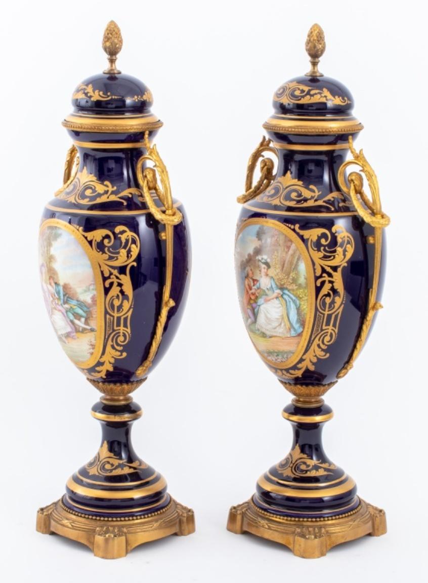 Pair of Sevres manner porcelain urns with gilt bronze mounted handles and bases, each with hand-painted courting scenes of lovers and landscapes upon cobalt blue grounds with gilt painted accents, the covers surmounted by pinecone form finials,