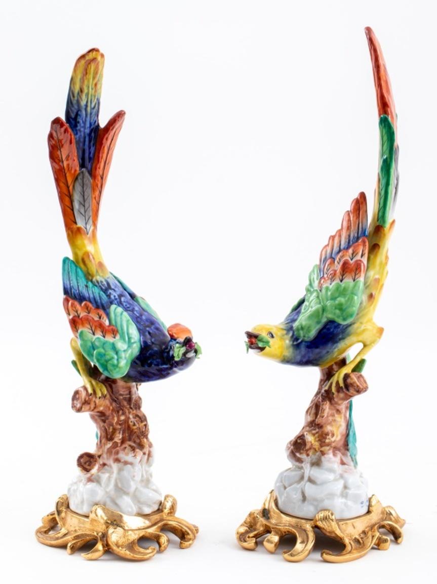 Pair of hand painted ceramic porcelain bird statue sculptures, each polychrome bird perched atop a tree trunk and holding fruit in beak, mounted on ormolu gilt bronze stands of scrolling foliate design, each bearing blue underglaze double Louis