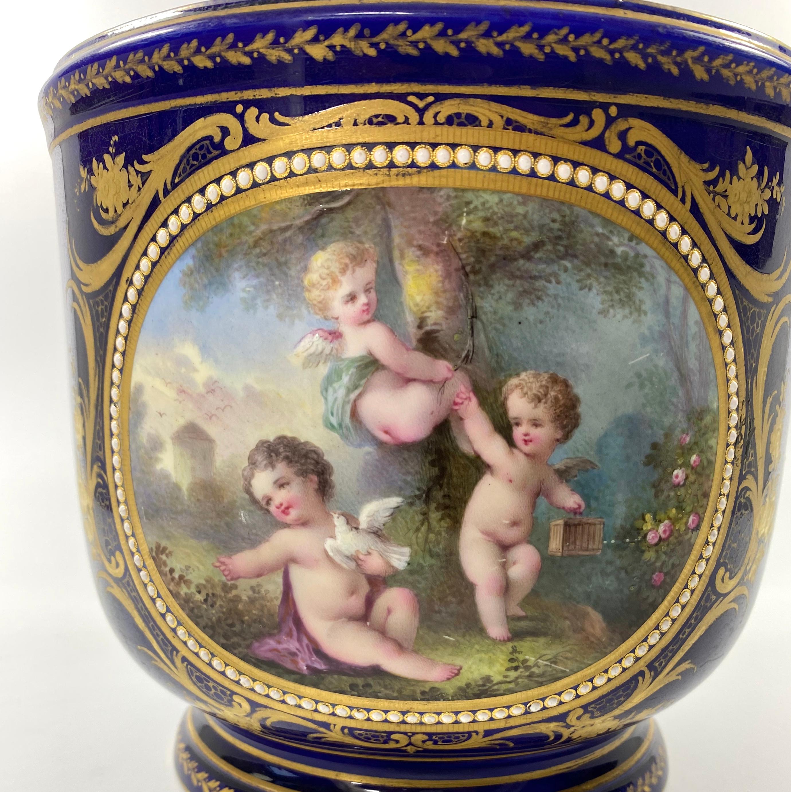 ‘Sevres’ style large porcelain ‘Jewelled’ cache pot, French, c. 1870. Finely painted with a panel of three Putti at play, in an extensive garden landscape, within an oval ‘jewelled’ border.
The reverse with a panel of doves, flying amongst symbols