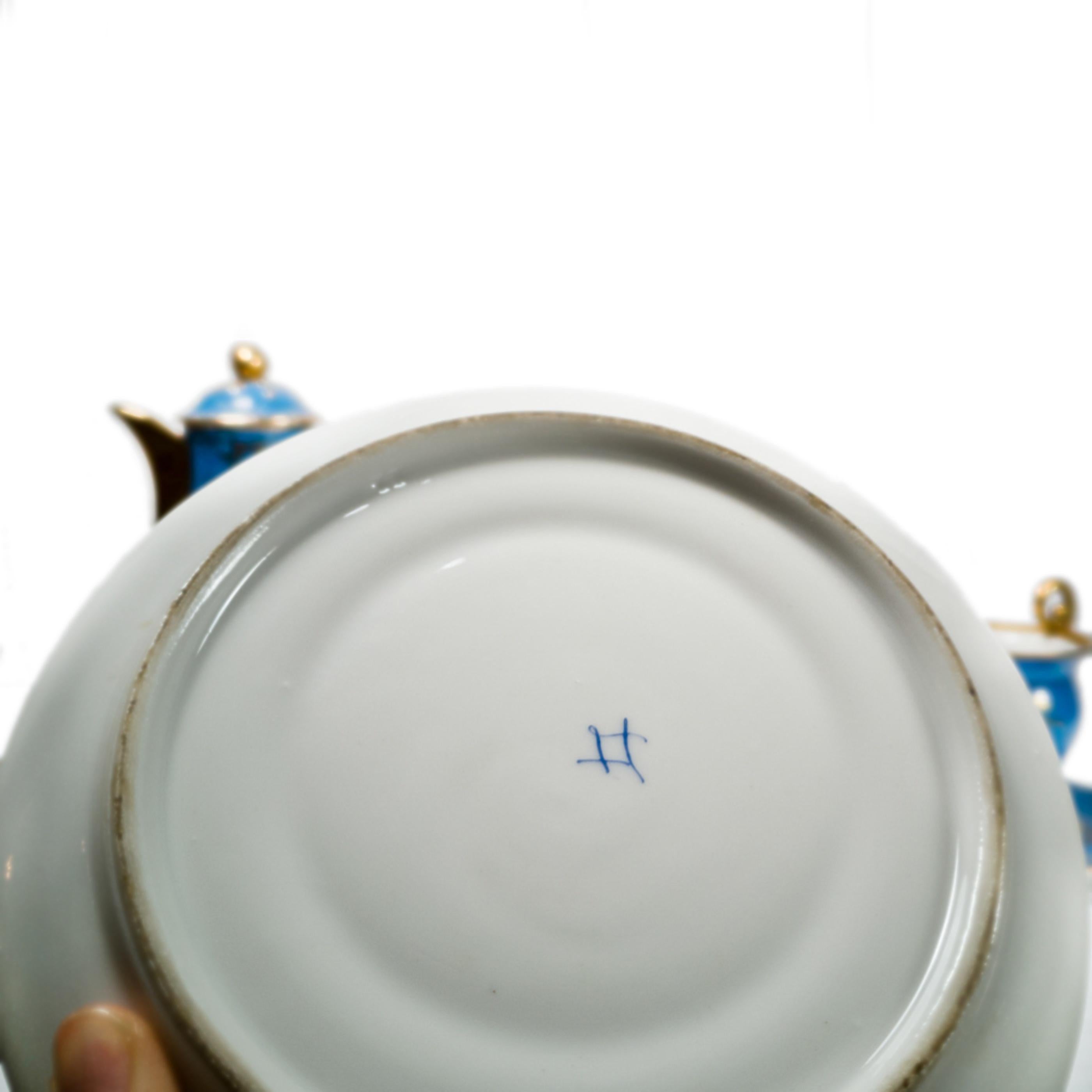 A set of exquisite blue and gold porcelain teaware, crafted in the esteemed Sèvres porcelain factory, mdel from 1764. The set includes a teapot and two cups and saucers, each piece adorned with intricate plant motifs and gold embellishments. The set
