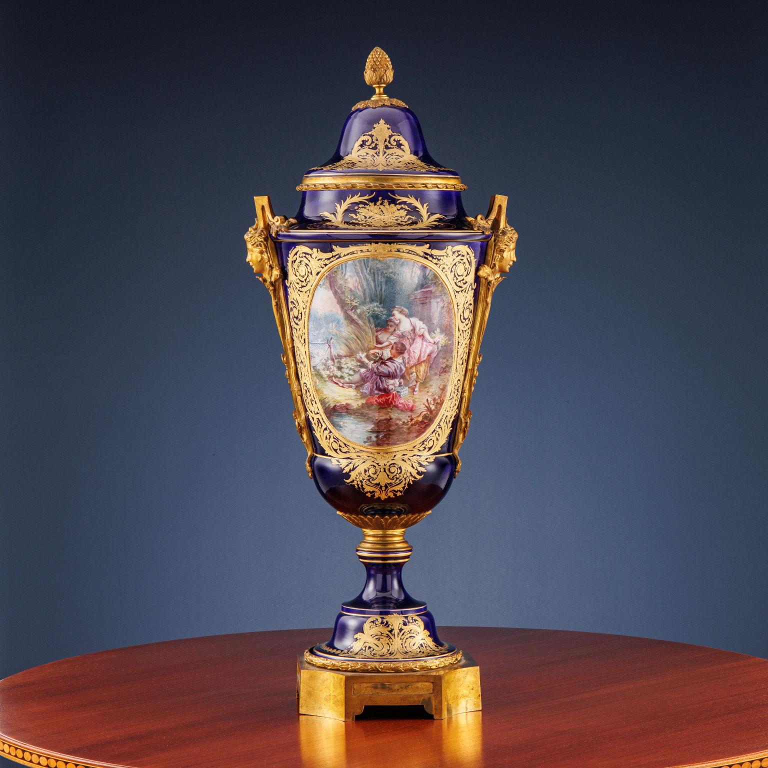 Sèvres porcelain urn vase in cobalt blue with gilded phytomorphic decorations and polychrome medallions, in one depicting a lake landscape, while in the other a bucolic scene; in the latter, he signs “Cottiret”. The polylobed base, the handles with