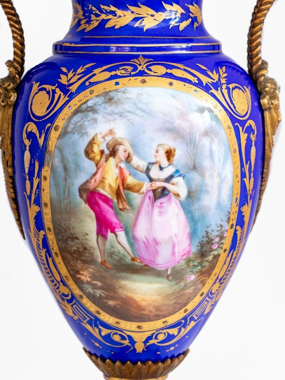 Sevres Style Blue Ground Porcelain Two Handle Covered Vase, early 20th century, with gilt metal mounts, painted with an oval scene of a dancing couple and a bouquet of flowers, within elaborately gilded borders. 18.5