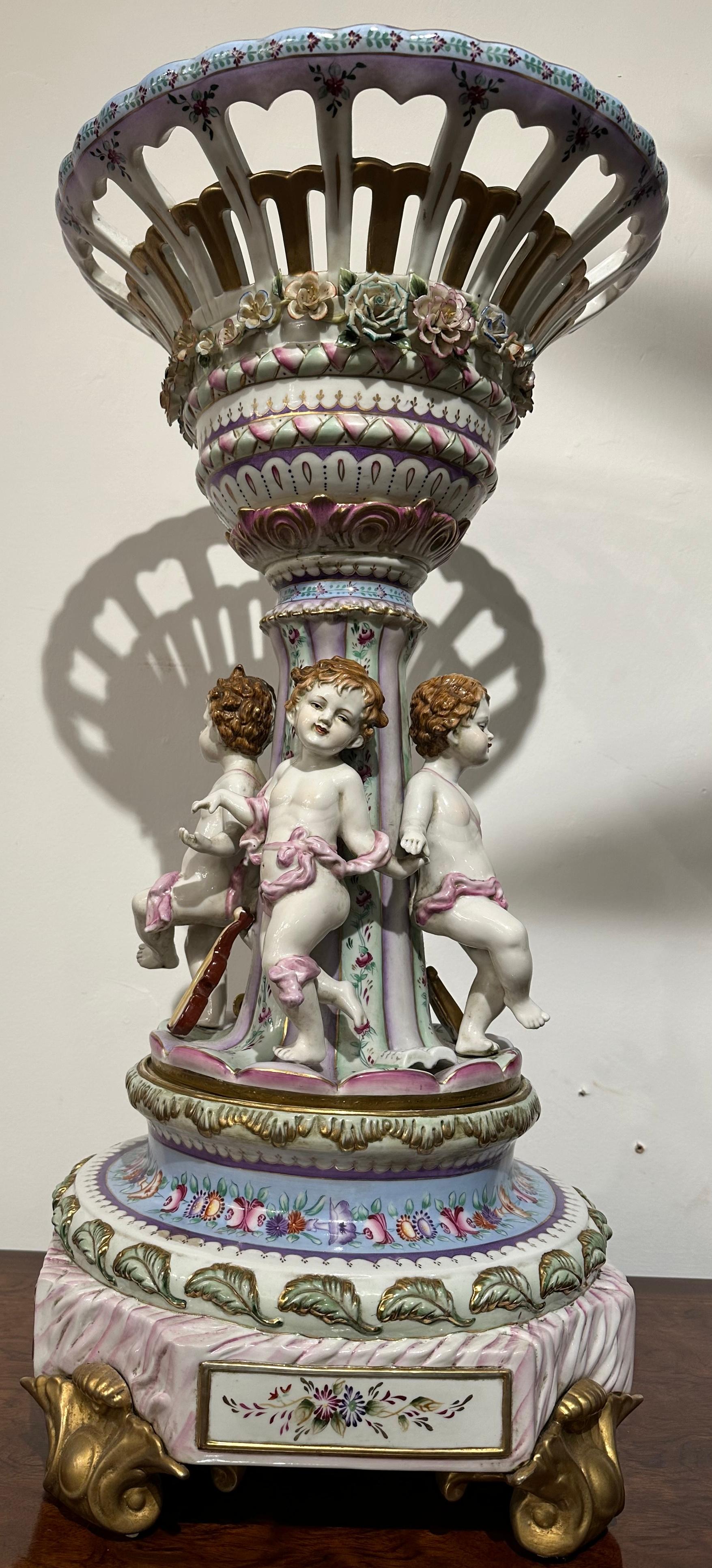 Ein äußerst dekorativer und sorgfältig gearbeiteter Tafelaufsatz aus Porzellan im Sèvres-Stil. Ein zarter, durchbrochener Korb, umkränzt von Rosen, sitzt auf einer Säule, die von Amoretten umgeben ist, auf einem kunstvoll bemalten, gestuften Sockel.