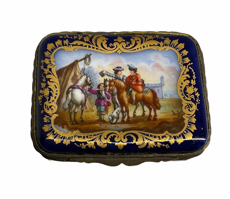 This is a Sevres style hand painted porcelain hinged small rectangular box. The background of the box is painted cobalt blue. It’s featuring in the front lid, a scene of some soldiers with their horses in their camp. In the back, you can see a