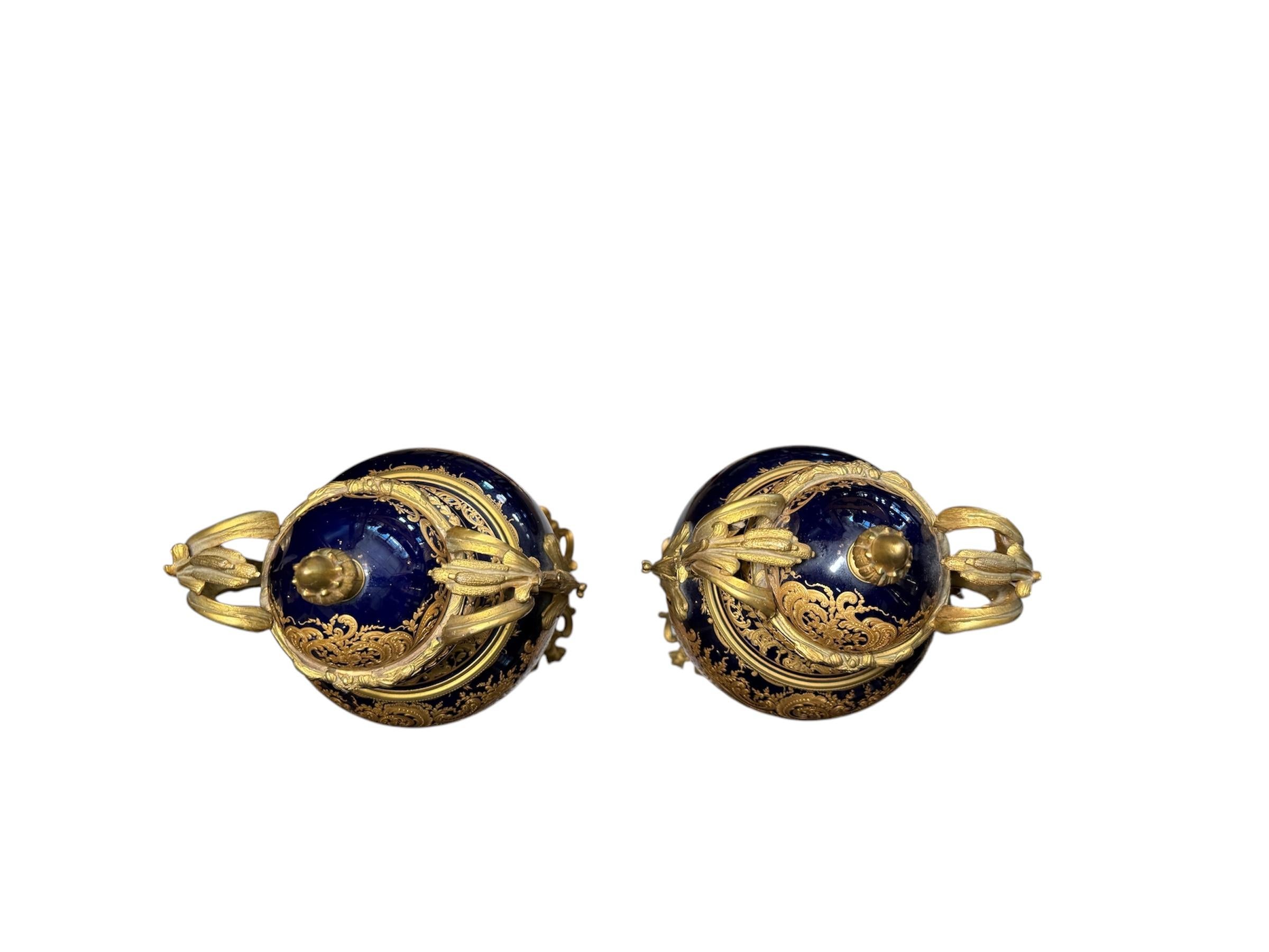 This is a Pair of Gilt Cobalt Blue Urns. Both Urns depict a cobalt blue background that is decorated in the center with a hand painted country side landscape enhanced by a maid playing with a Cupid. The scenes are framed by a design of gilt garland