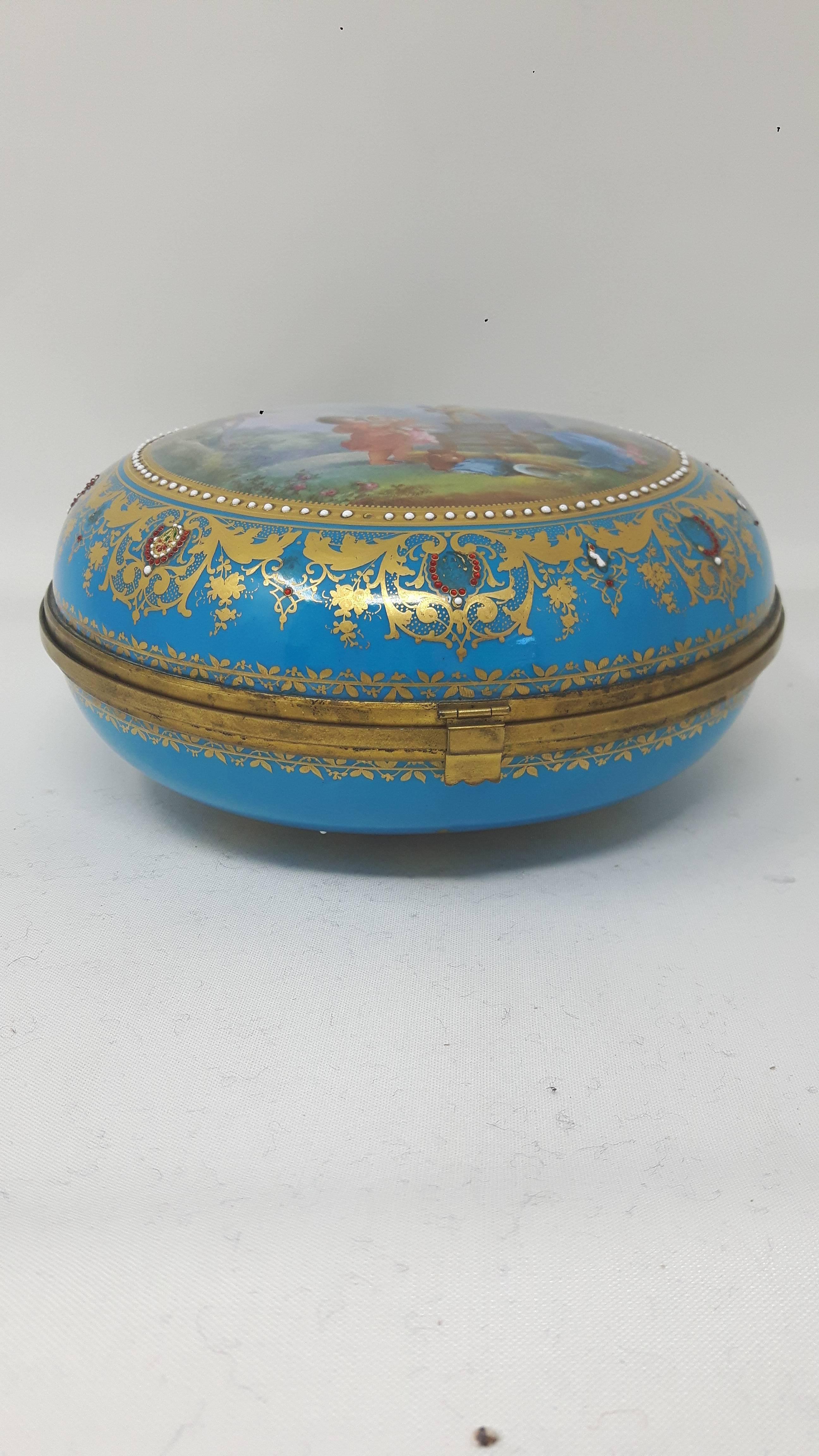 A Sèvres style Paris porcelain round box with ormolu mounts, hand-painted with a romantic scene of two children playing with a bird. The borders are elaborately decorated with gilded friezes and enamelled jewels, as is the bottom of the bo.