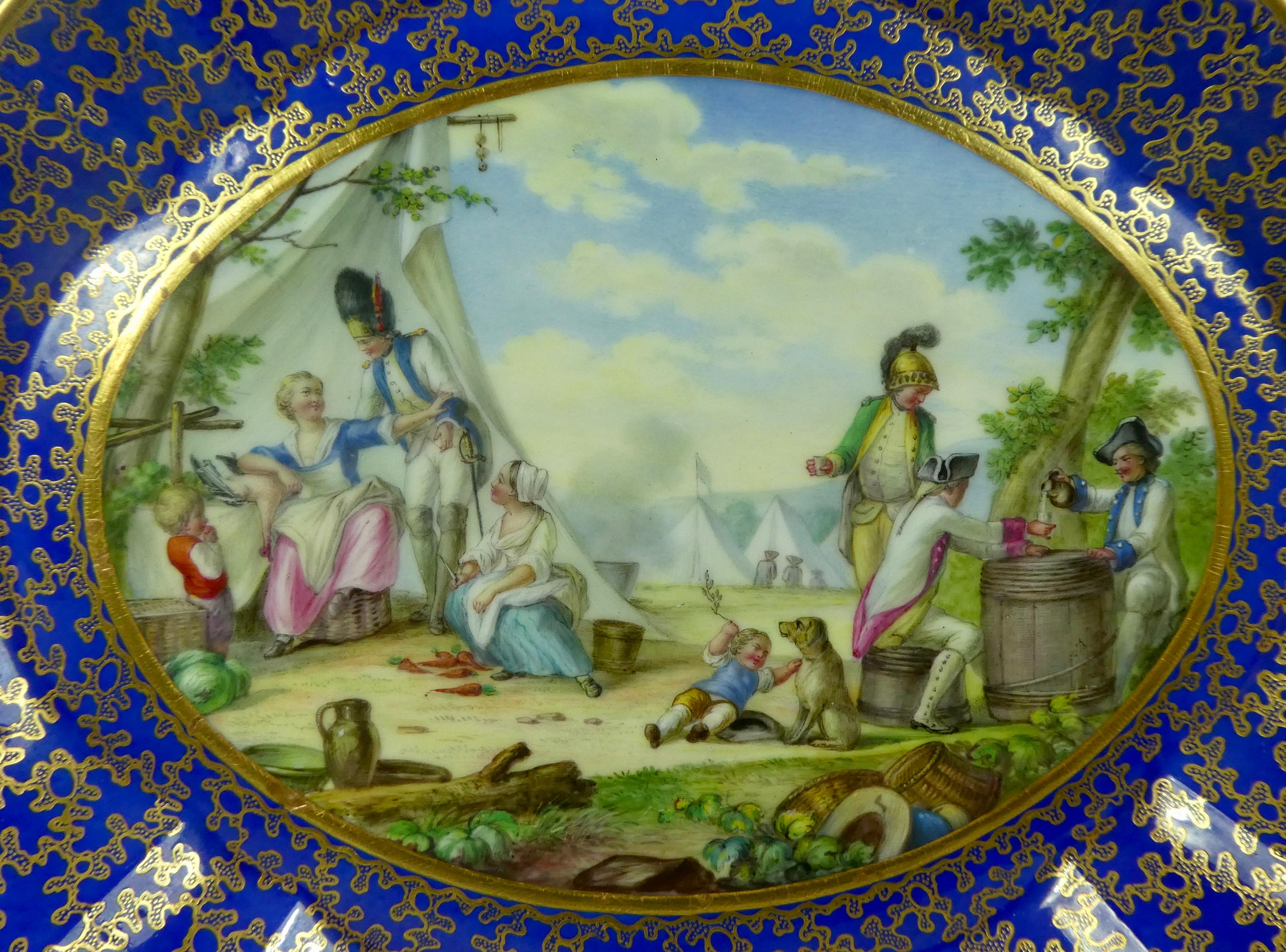 A large French serves style porcelain oval dish, circa 1850. The moulded dish, finely painted to the center with a scene of soldiers and civilians relaxing in a military encampment, dyer a painting by Teniers. Within a heavily gilded cobalt blue