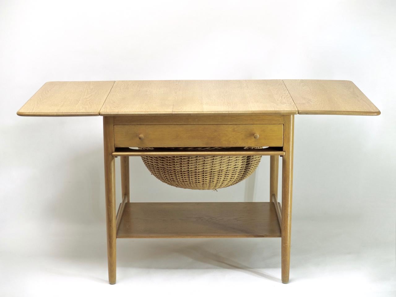 Sewing table, side table by Hans J. Wegner for Andreas Tuck made of oak with a pronounced wood grain from the 1950s. With 2 table extensions that can be folded up, a pull-out wicker basket, a drawer with holders for yarn, needles and accessories and