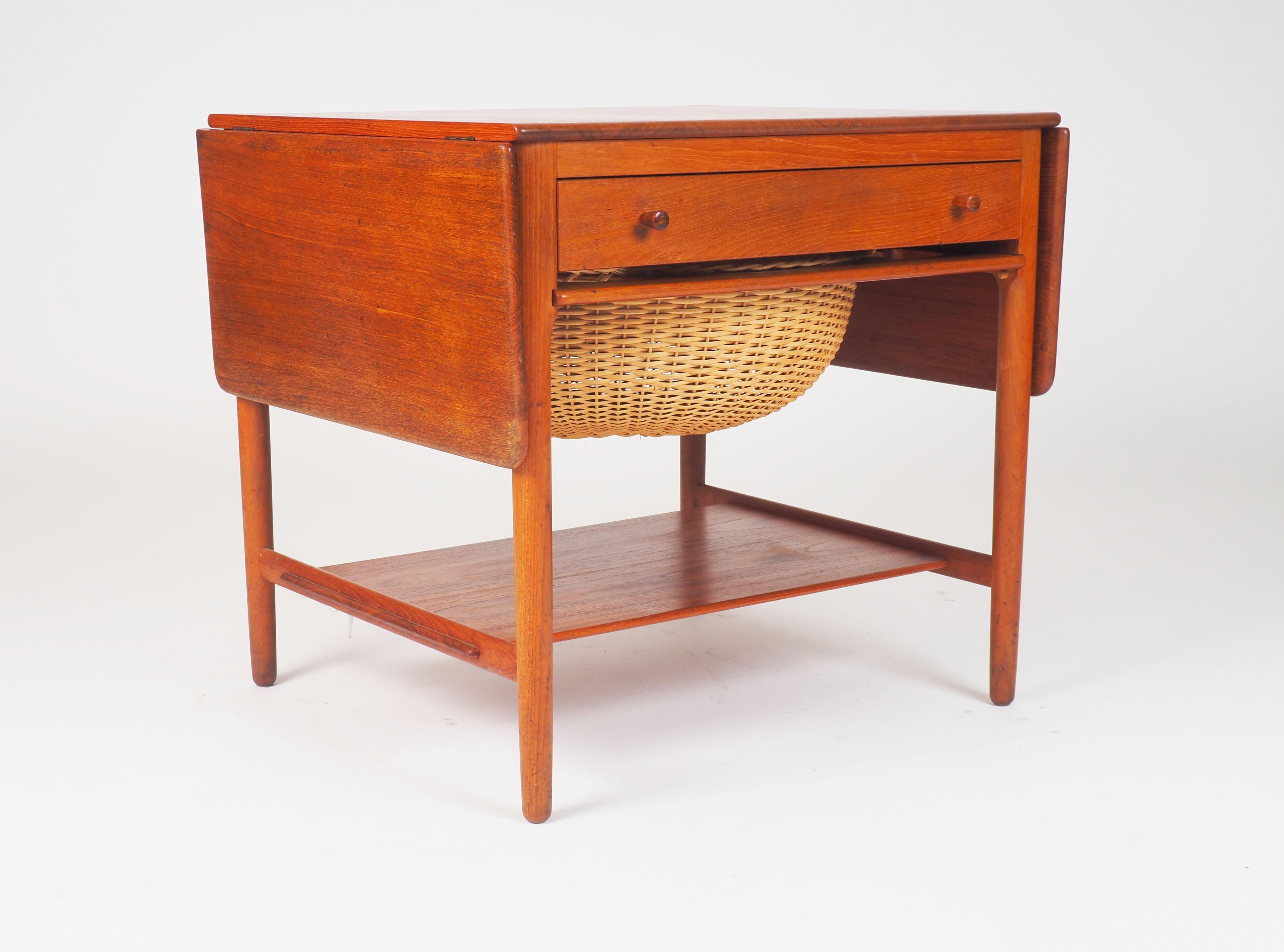 Sewing table in solid teak and oak, designed by Hans J Wegner and made by cabinetmaker Andreas Tuck. The table has two drop-leafs, a front drawer with several compartments and a handwoven rattan basket for knitting’s.