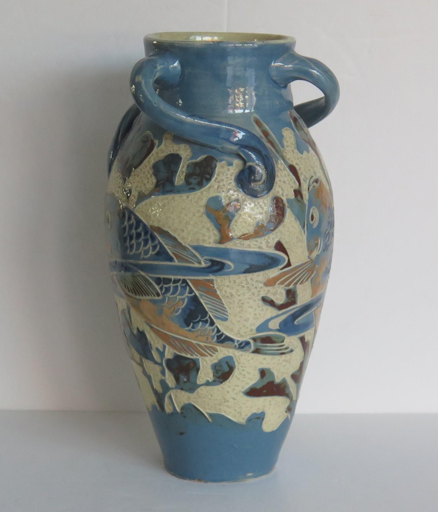 Il s'agit d'un bon vase en poterie d'art, avec trois anses angulaires, toutes modelées à la main selon la méthode du sgraffite, avec une décoration de poissons, fabriqué à la main par C H Brannam's, en Angleterre, et datant de la fin de la période