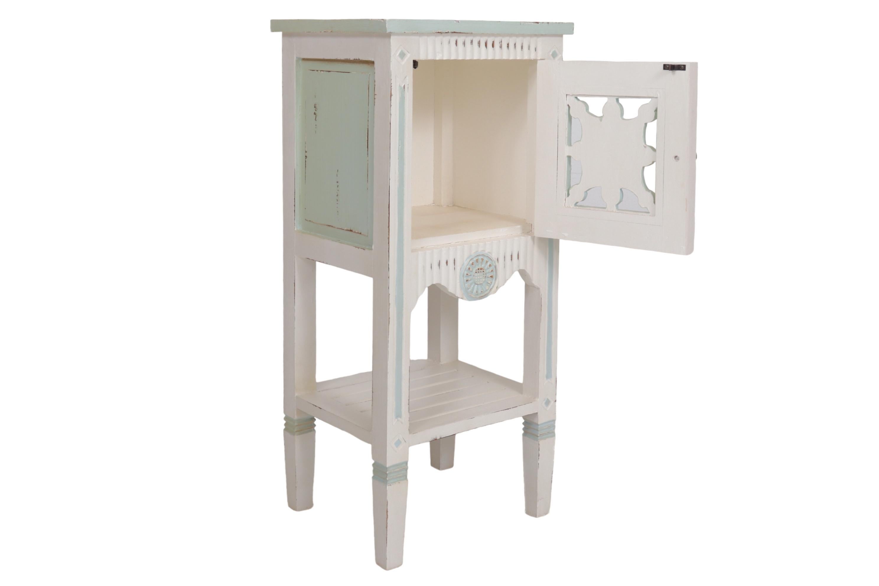 A shabby chic side table made of solid wood and painted in white with teal details. The cabinet door is pierced and decorated with a simply carved sunflower motif that repeats on the reeded skirt below. Straight square legs support a slatted shelf