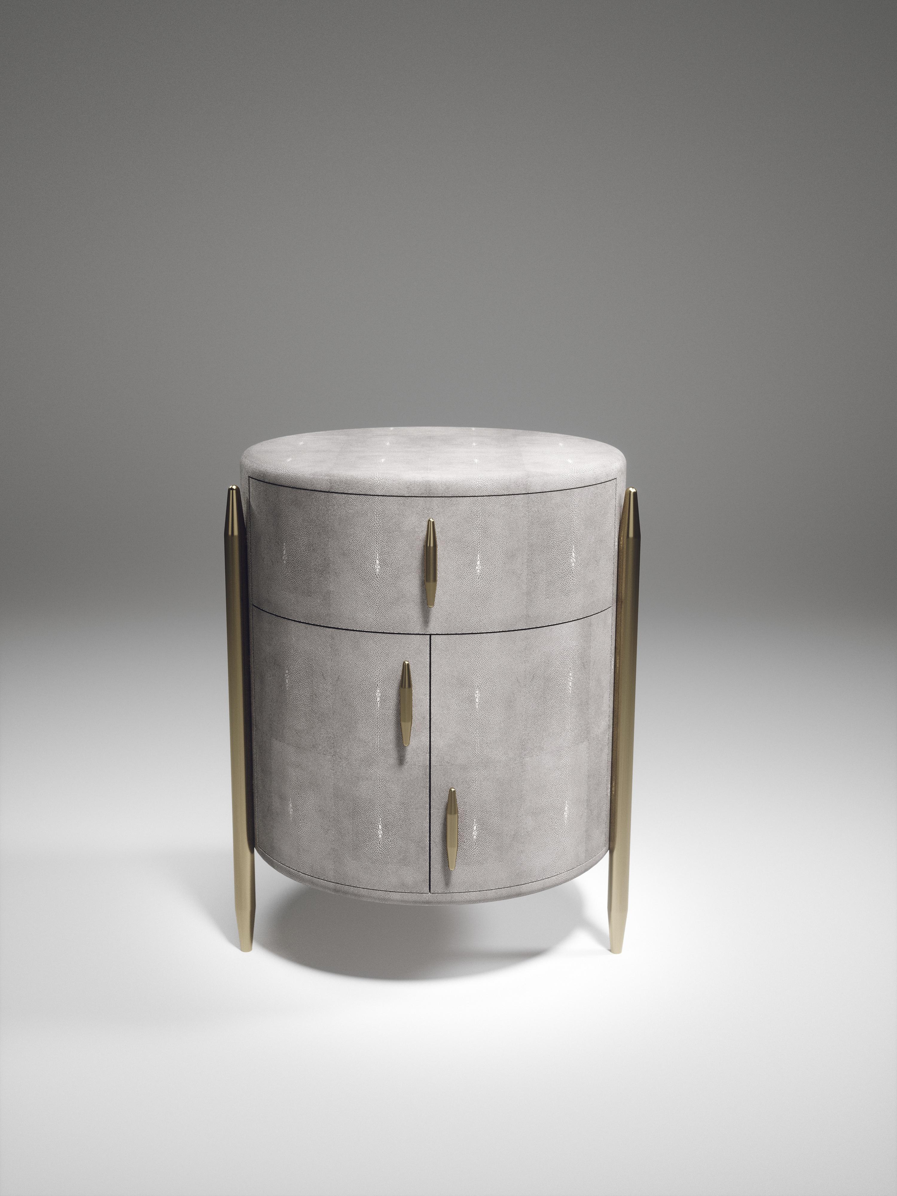 The Dandy round bedside table by Kifu Paris is an elegant and a luxurious home accent, inlaid in light grey shagreen with bronze-patina brass details. This piece includes 1 drawer total and a cabinet below; the interiors are inlaid in gemelina wood