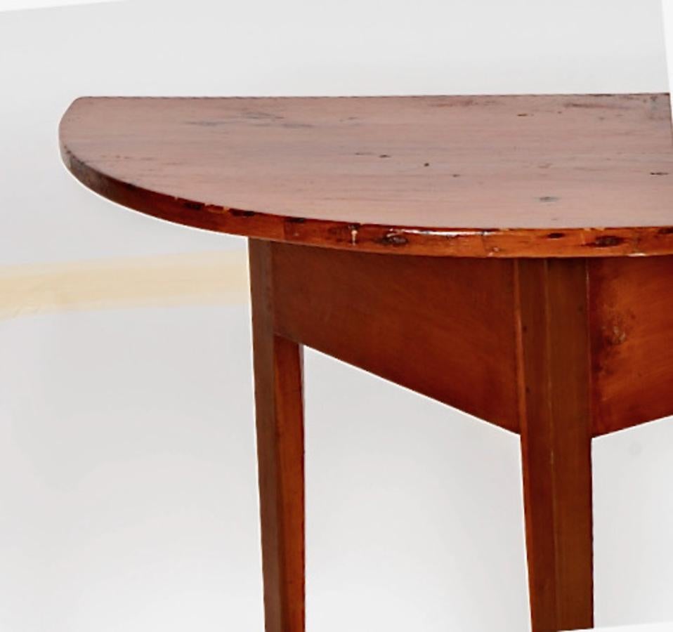 This is a charming mid-19th century pine demi-lune table that has been attributed to Shaker fabrication. The demi-lune measures a generous 40 inches wide and features a deep overhang on the triangular frame. The delicate legs narrow at the toes and