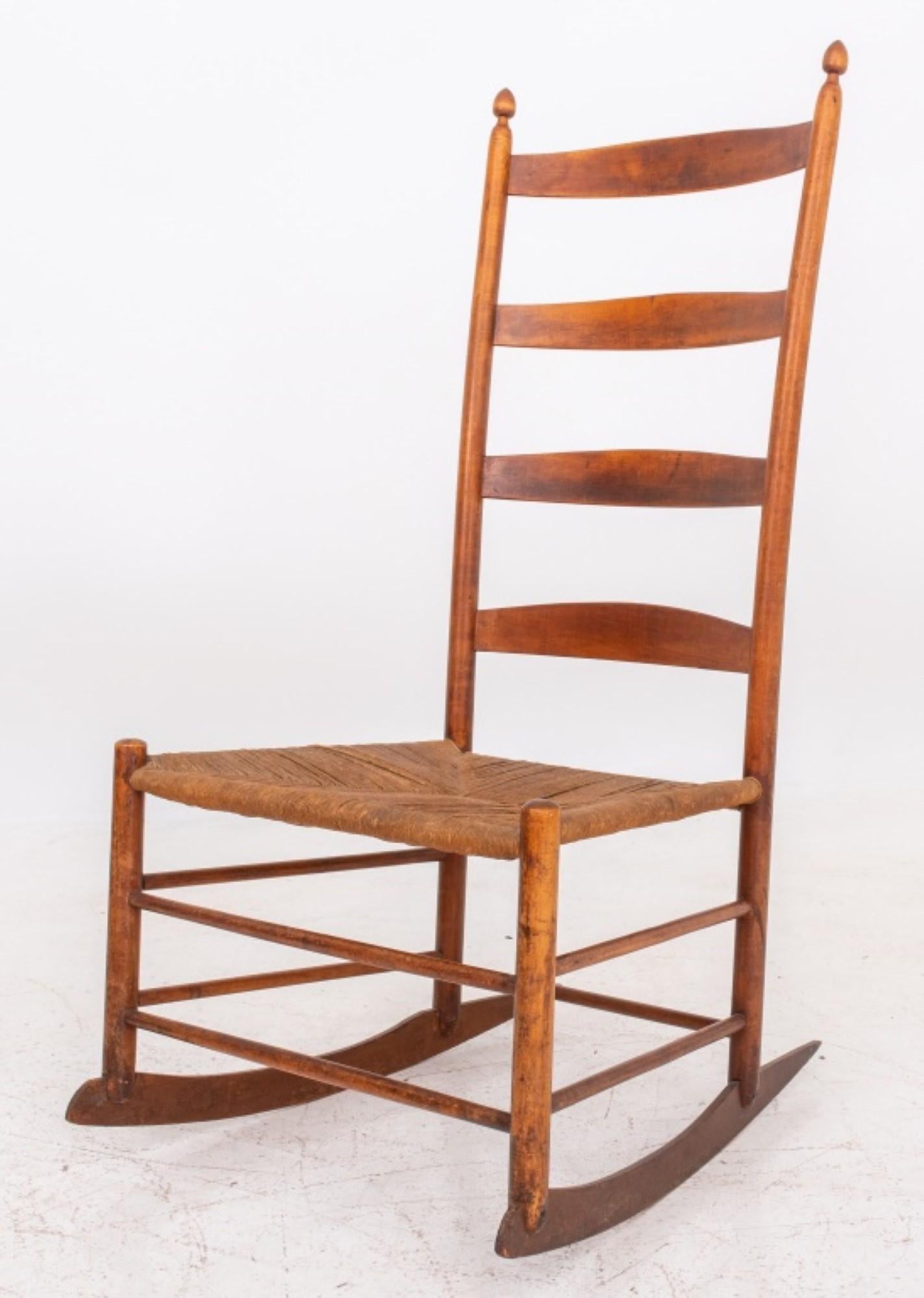 Shaker rocking chair in fruitwood. Here are the details:

Style: Shaker
Material: Fruitwood
Features:
Finials above four ladder back splats
Rush-woven seat
Four legs conjoined by stretchers
Legs above rockers (possibly later reinforced with