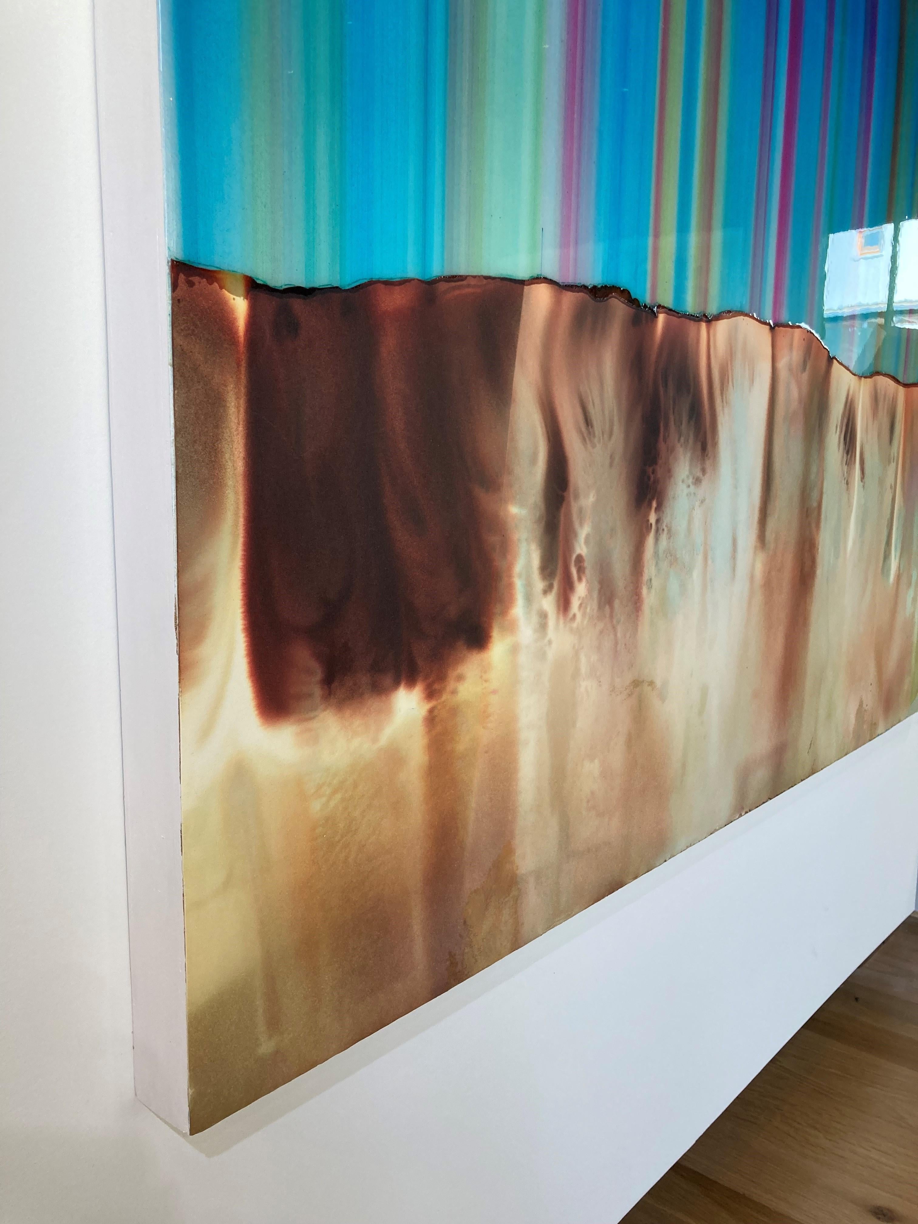 Striations of bright blue, teal green and pink in vertical stripes create an abstract horizon line suggesting a vibrant striped sky over a vast mountain range in golden brown with hints of bright olive green, while layers of resin on the top half
