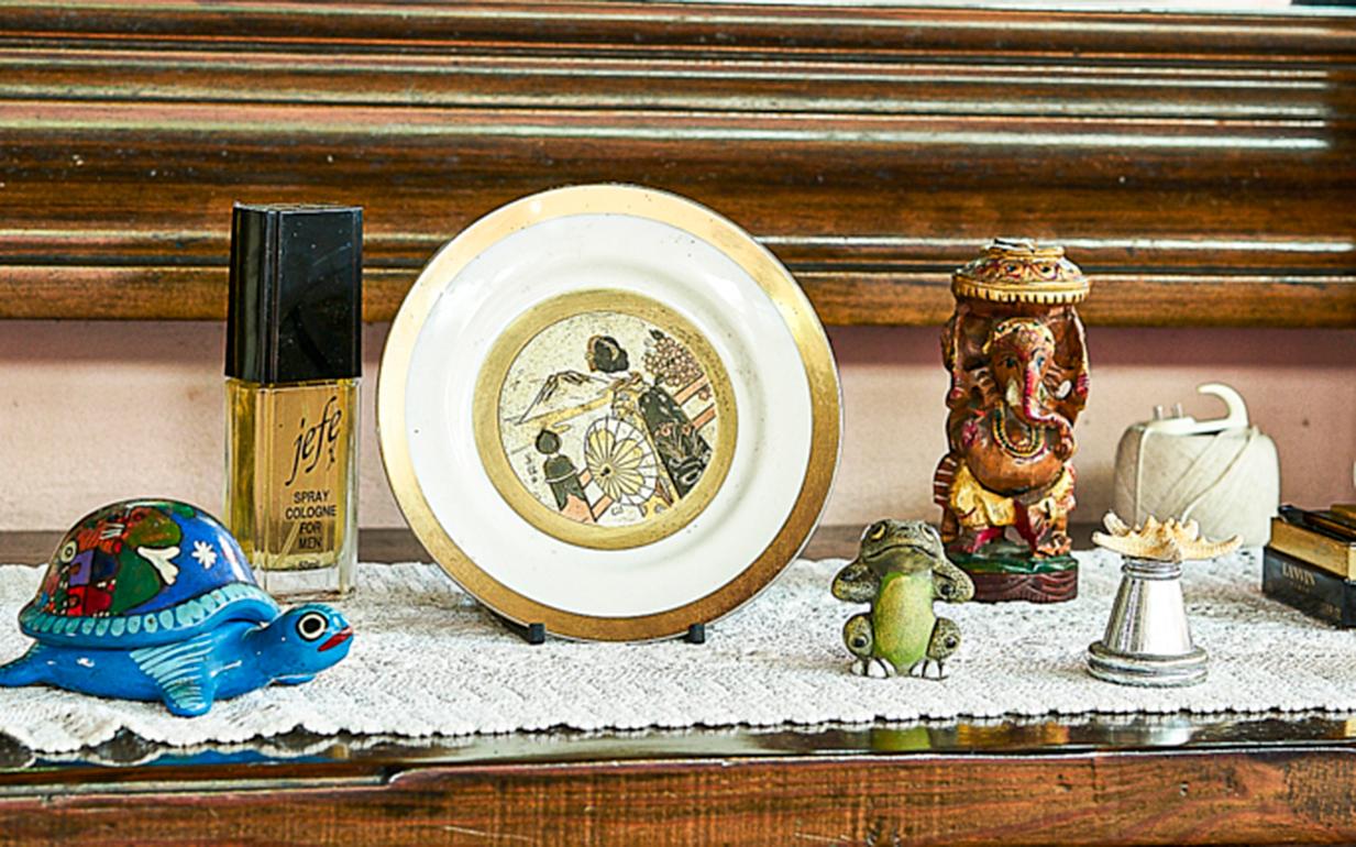 Eine Kommode aus dunklem Holz, eine Sammlung von Gegenständen - Familienfotos, Schildkrötenfiguren aus Porzellan, ein Porzellanteller, eine Flasche Parfüm - bedeutungsvolle Gegenstände, die eine Geschichte erzählen. Dies ist das trinidadische Haus,