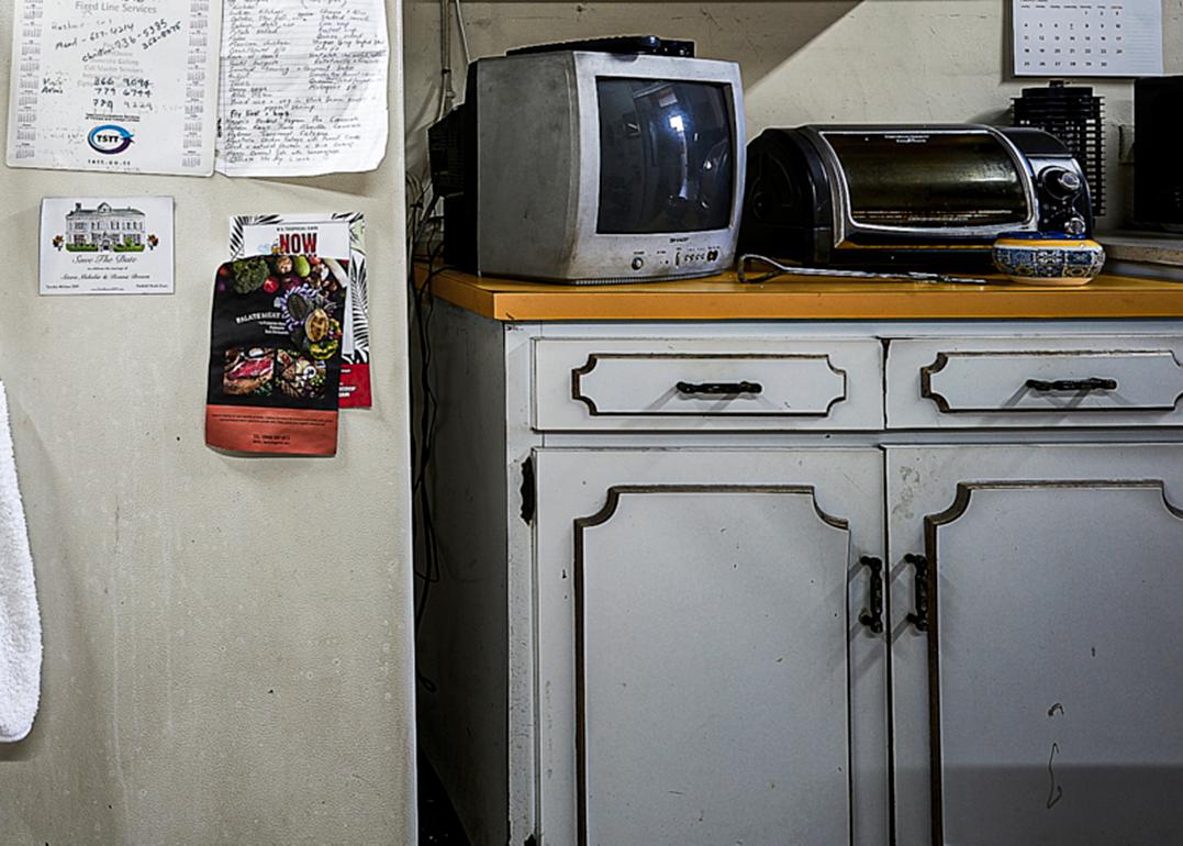 Shani Mootoos Fotografien sind Porträts eines Lebens, die zugleich intim und einnehmend sind. Dieses Bild einer Küche - die abgenutzten weißen Schränke, der mit Notizen und Fotos bedeckte Kühlschrank, der Toaster und ein alter Fernseher auf dem