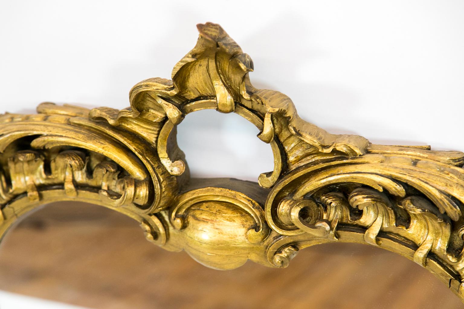Shaped gilt Rococo mirror is framed with stylized waves carved in high relief. It has a flame crest and a fluted, voluted base.
