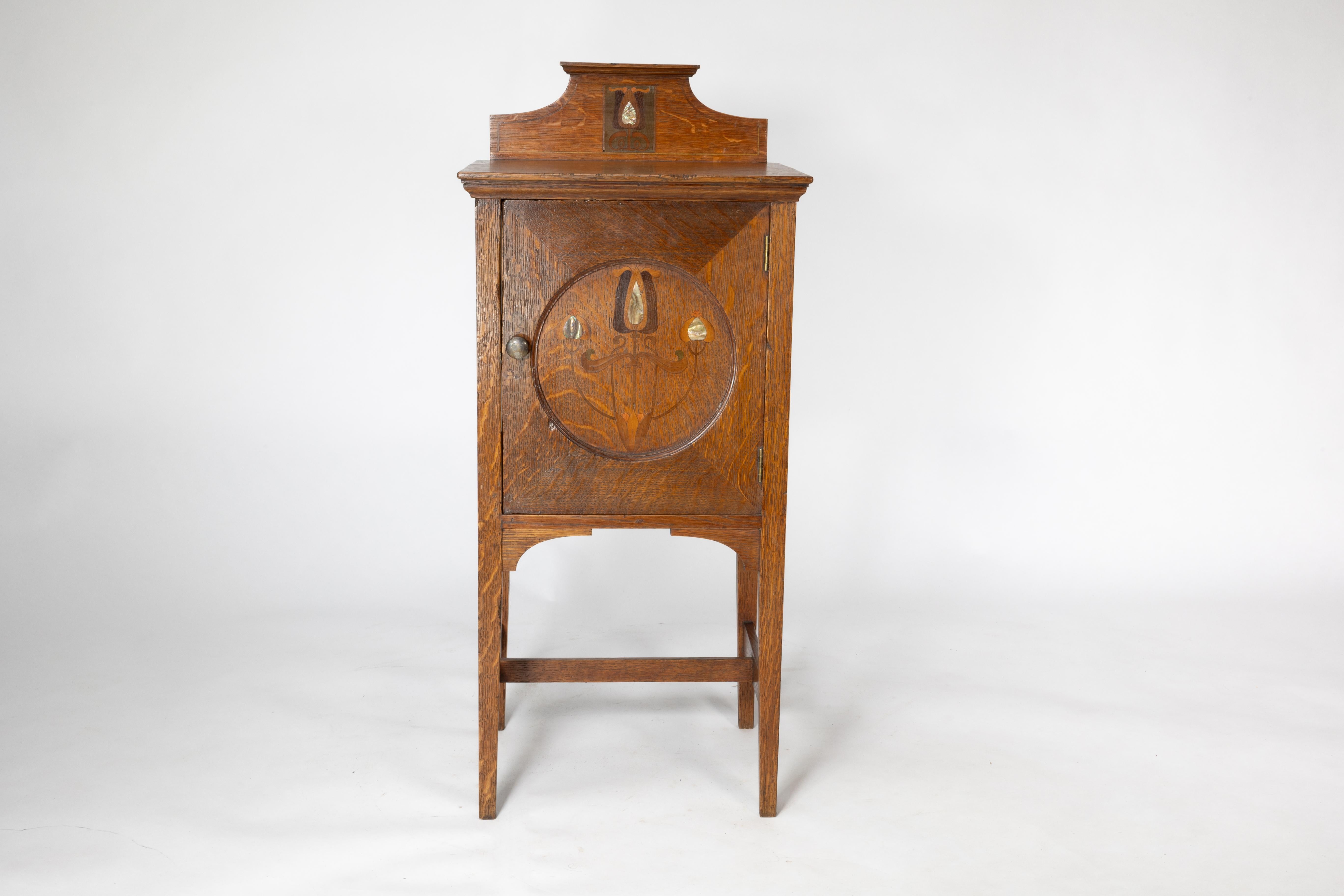 Shapland and Petter. An Arts and Crafts oak bedside cabinet with stylized floral inlaid decoration in the form of Albalone flower heads.