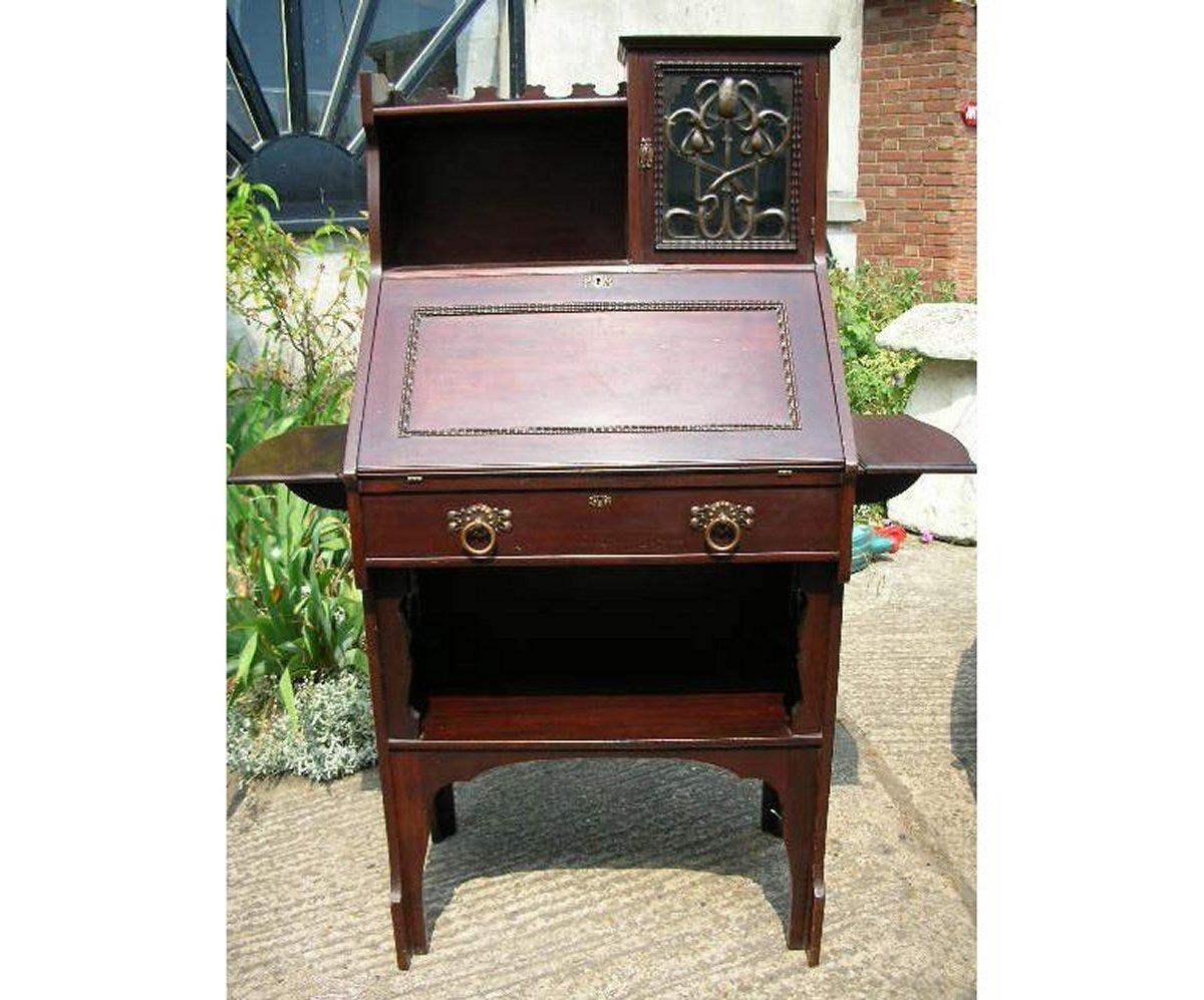 An Arts and Crafts fold down mahogany desk by Shapland and Petter. Comprising of a cupboard to the top right, it's glazed door with a copper stylised floral panel; upper and lower bookshelves to it's left; a full size work surface which folds down