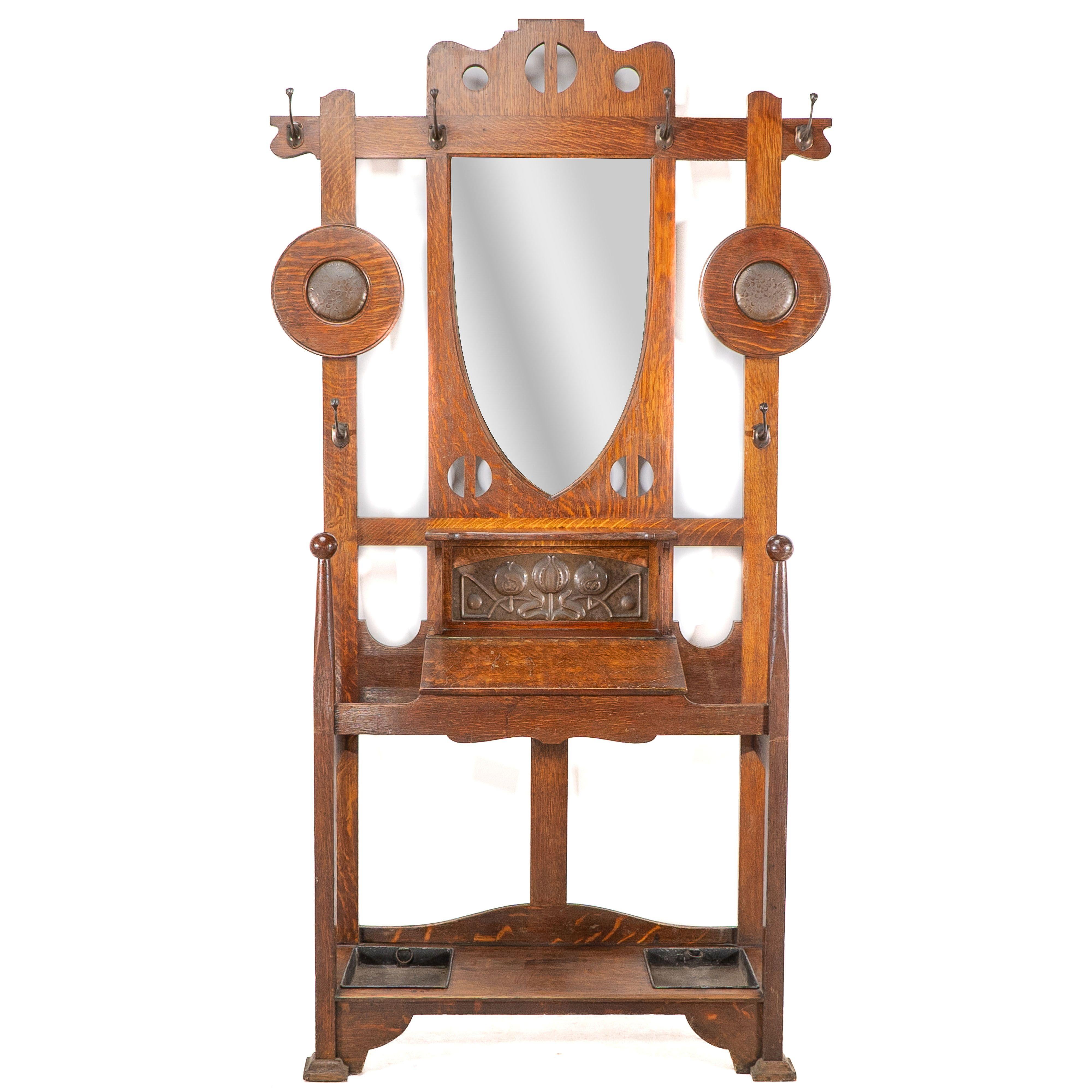 A top quality Arts and Crafts oak Hallstand by Shapland and Petter with bevelled mirror flanked by a circular copper disc with hat and coat hooks, stylised cut-outs and stylised floral copper panel and glove box, the front sculptured uprights capped