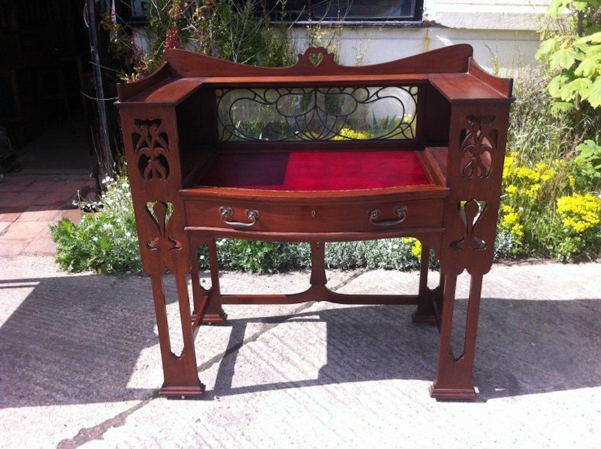 Shapland & Petter. A wonderful Arts & Crafts mahogany writing desk with a pierced heart to the top, shaped back and sides, with upper three quarter shelf and a stylized floral leaded glass back below. A period style leather writing area with hand