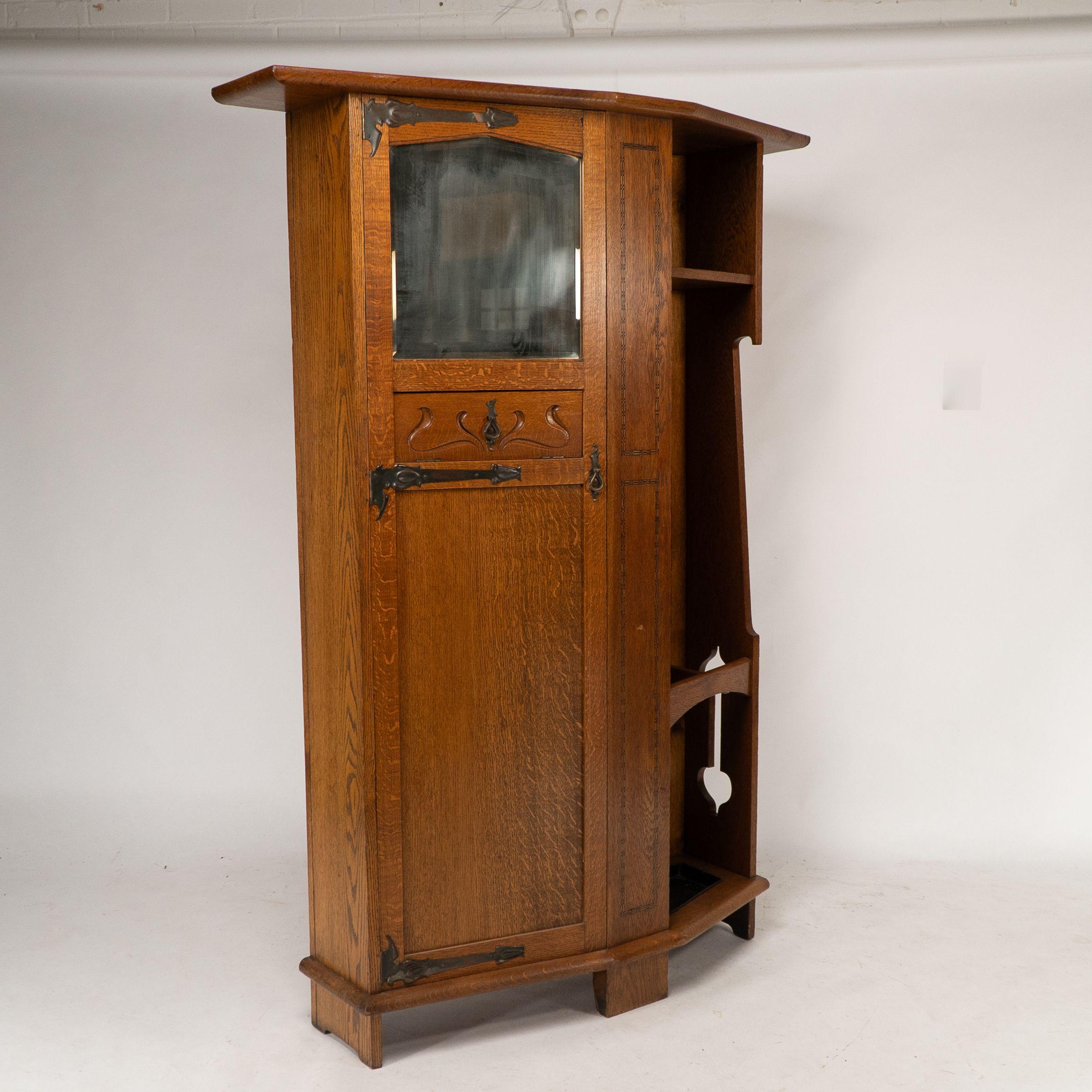 An Arts and Crafts oak hall cupboard attributed to Shapland and Petter with a flaring cornice perfect to pop hats on, an upper bevelled mirror and an unusual half revolving drawer for gloves and keys inset into the middle of the left hand door with