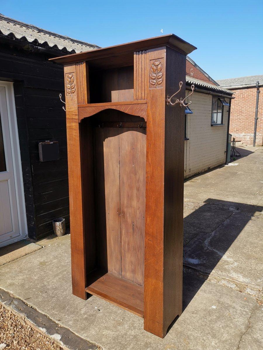 Shapland and Petter. An Arts & Crafts oak hall cupboard in the style of M.H. Baillie Scott, of tapering Architectural form. The flaring cornice with an open upper hat cupboard, flanked by stylized floral carvings. The open hanging coat space has