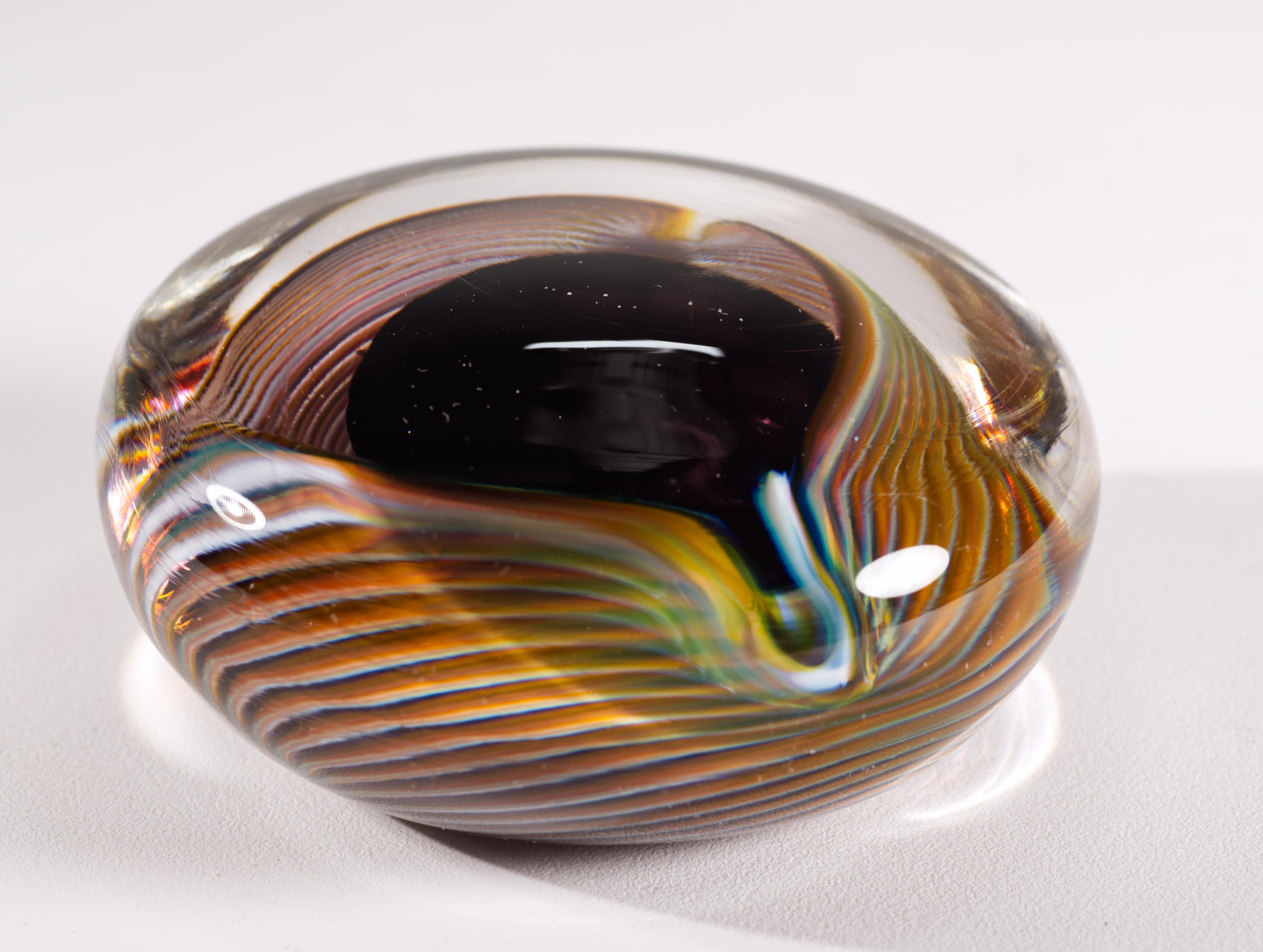  Le presse-papier en verre d'art vintage présente un design Op-Art avec une structure circulaire en verre rouge bourgogne foncé, presque noir, entourée d'un ruban de verre rayé dans les couleurs bleu, blanc et orange brûlé ; l'élément décoratif