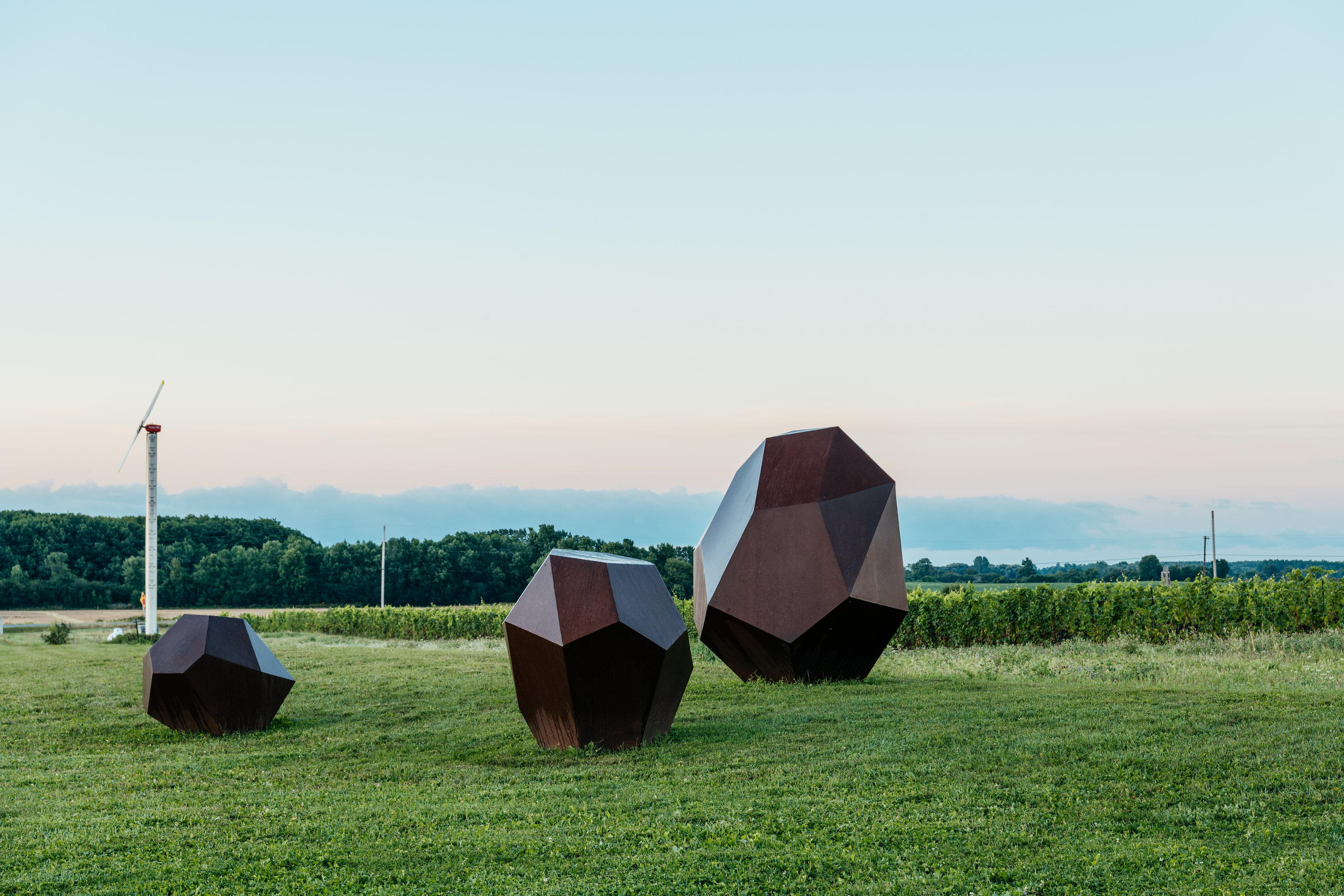 This large scale outdoor installation by Shayne Dark consists of three hollow, polygonal corten steel boulders called Drop Stones that have a rusted, weathered patina. The works represent erratics, geometric stones shaped and carried often hundreds