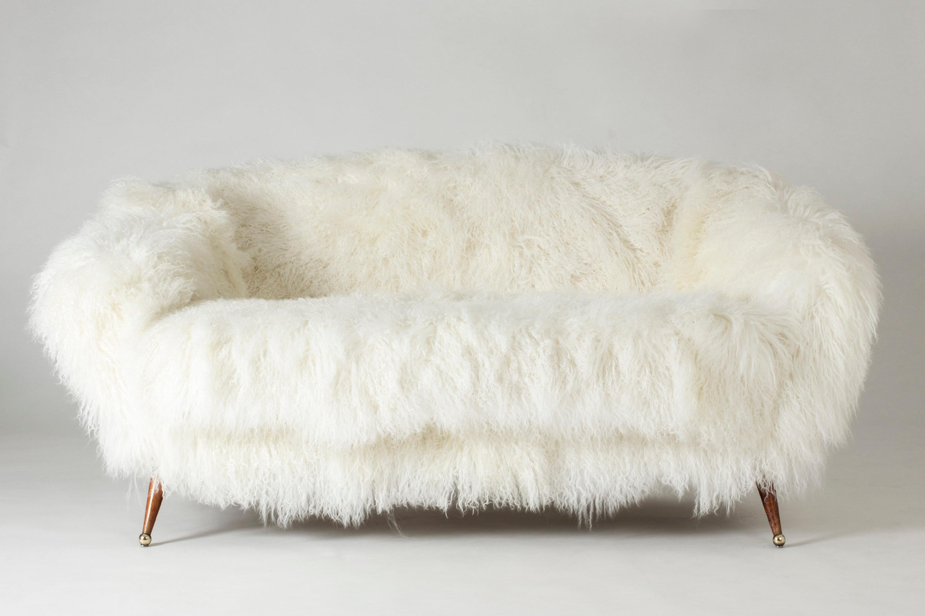 Stunning “Tellus” sofa by Folke Jansson, upholstered with white, Tibetan sheepskin. Rounded, wide and low design with slender legs with brass balls as feet.