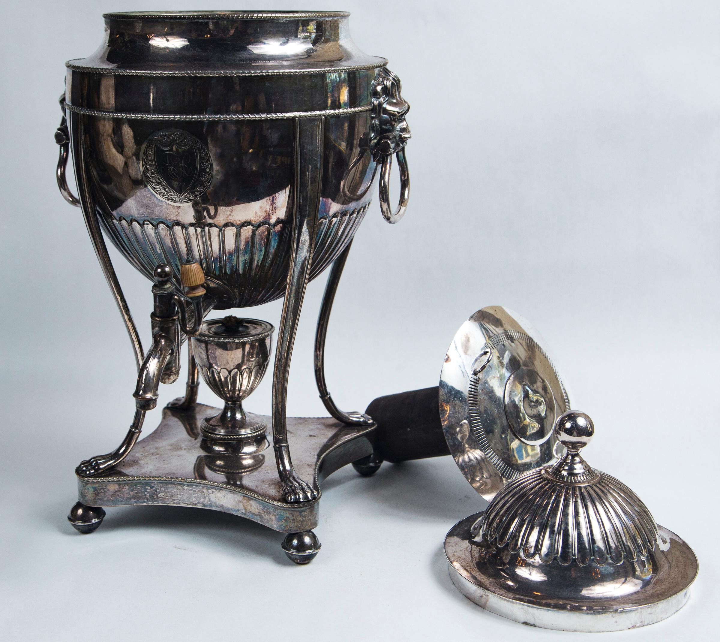In classical George III form. Complete with burner and cylinder for hot coal or stones. Concave base on ball feet support the urn that has legs with paw feet. Lion mask ring handles. A shield shaped with monogram and date of 1790 within a circle.
  