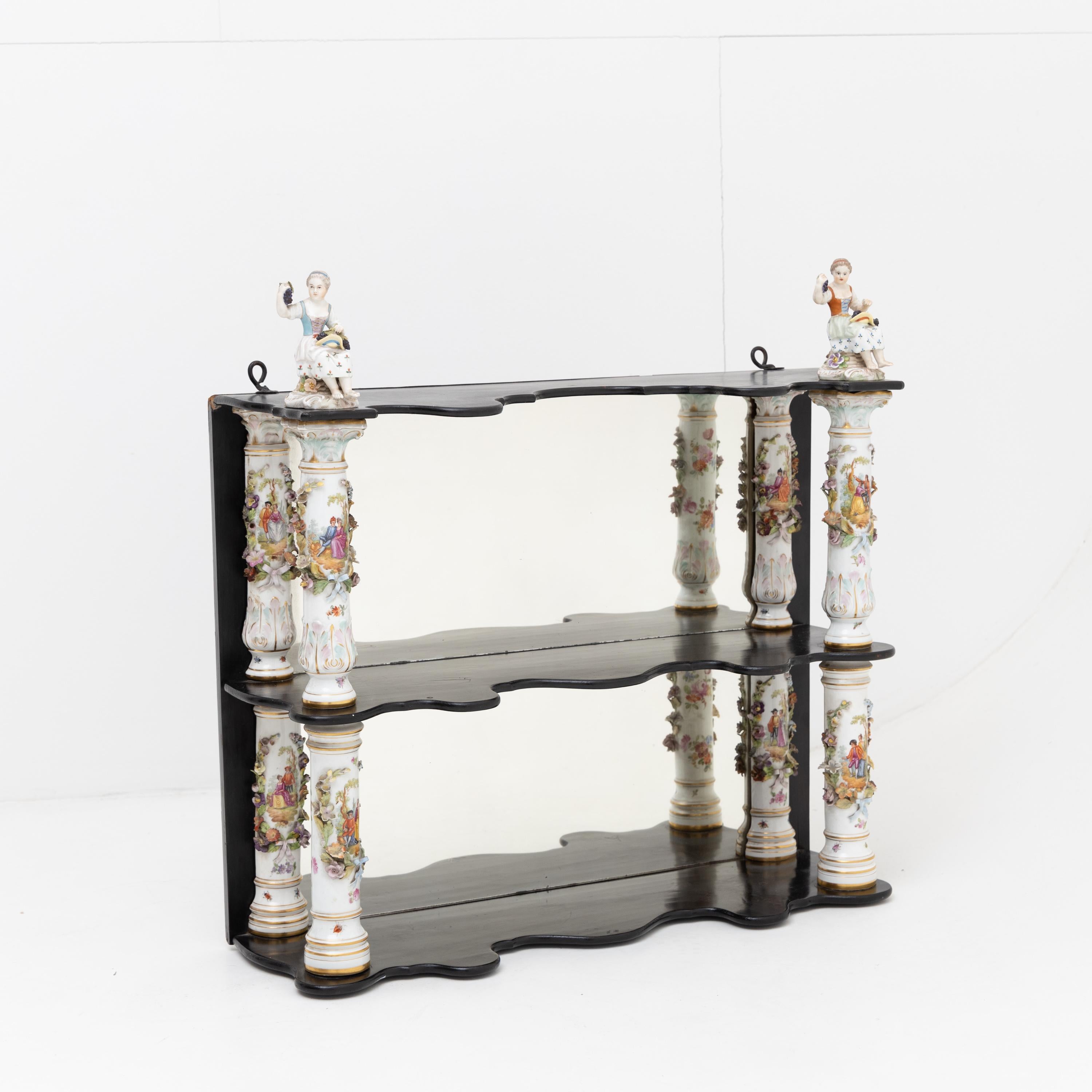 Small, ebonized wall shelf with polychrome porcelain columns and applied floral decoration and gallant figural scenes. The shelves are wavy, and the rear wall is mirrored. Small figurines of two girls with grapes sit on top of the shelf; marked Carl
