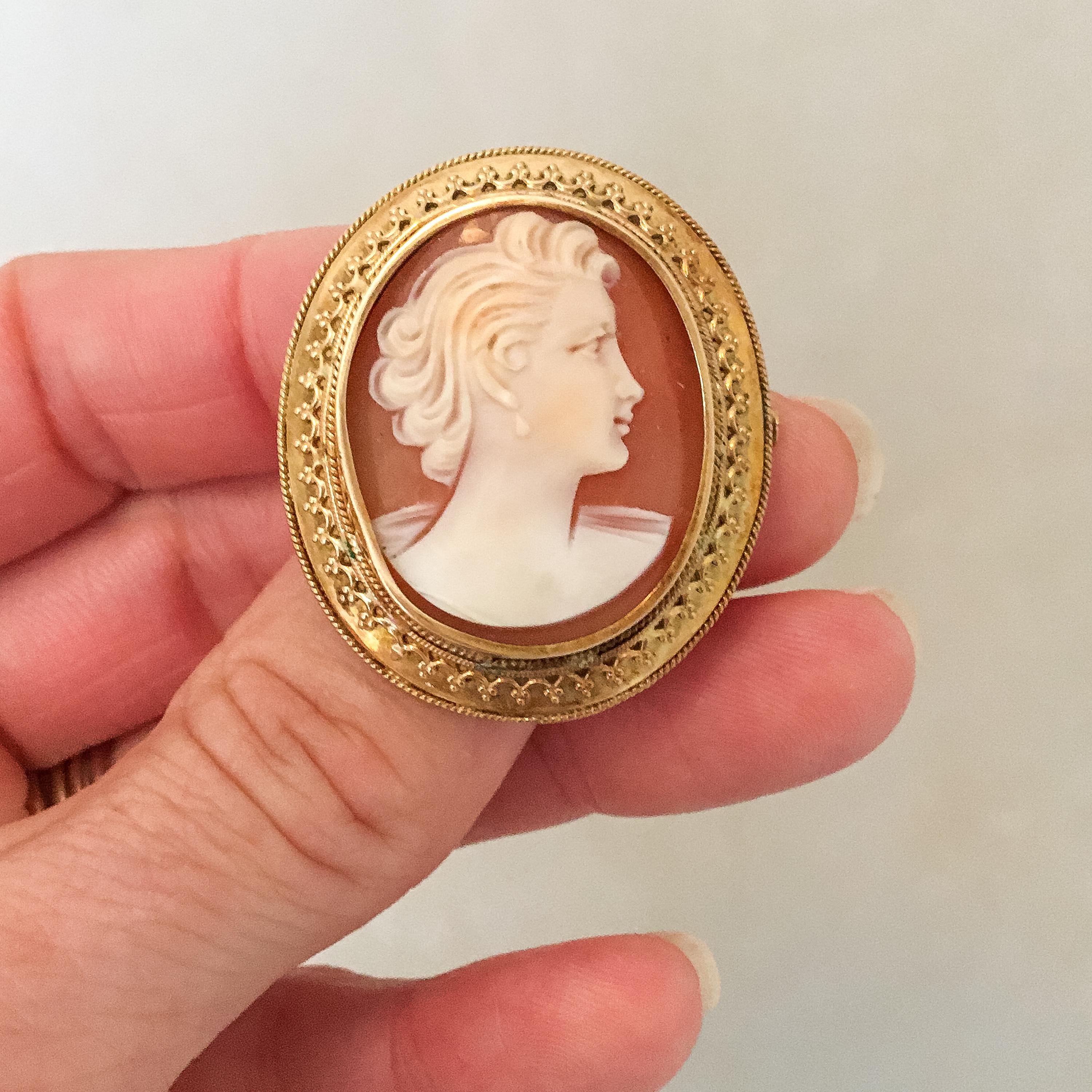 A vintage shell cameo brooch set in a 14 karat gold frame. The shell has a beautiful hue of pinkish and orangish colors and a white relief, and lies in a raised gold rim. The shell is carved into this female silhouette design while it is set in a