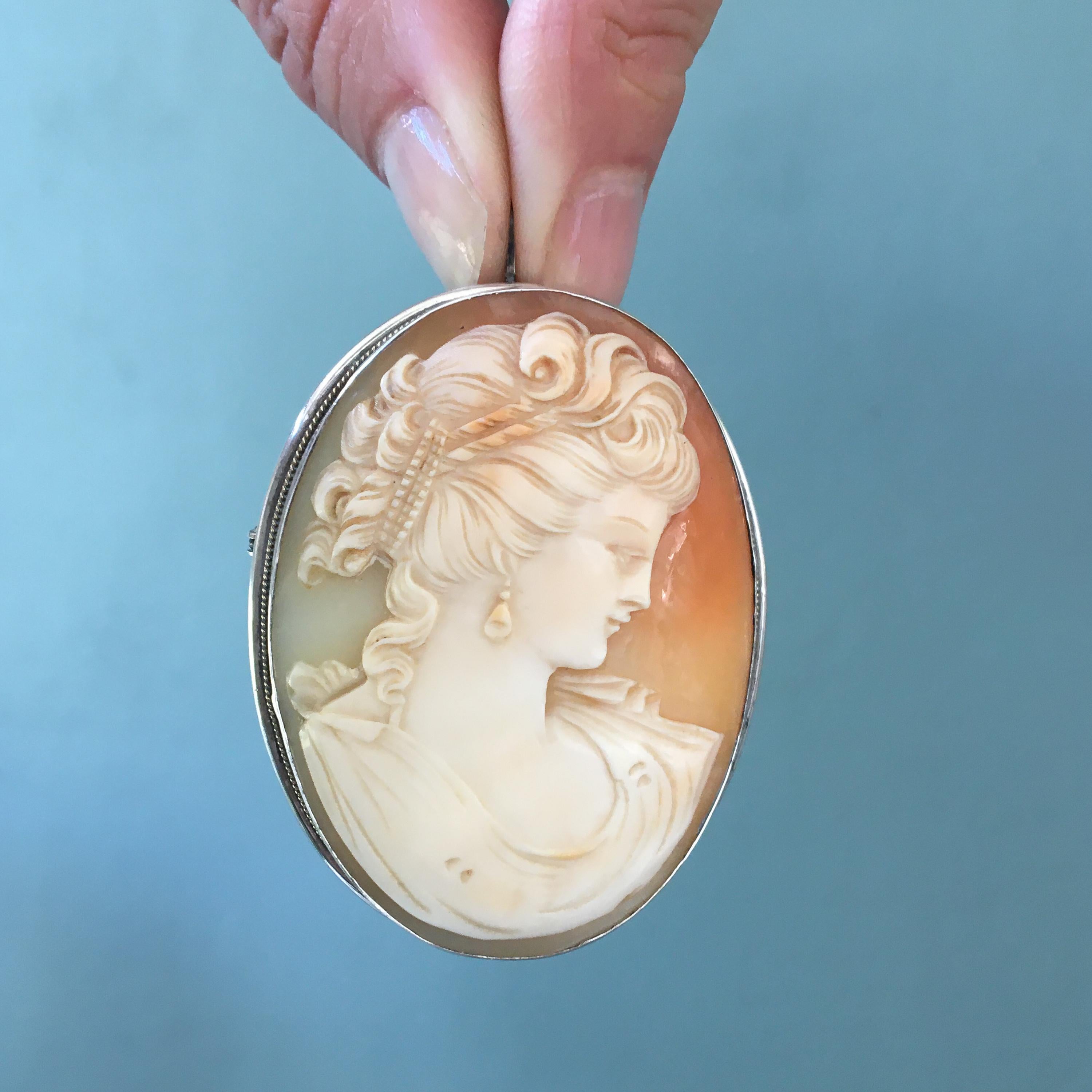 A large shell cameo brooch set in a silver frame. The cameo is very detailed into a carved female silhouette. The woman radiates beauty. Her curly hair is beautifully detailed with a ribbon in her hair and wearing drop earrings. The carved shell is