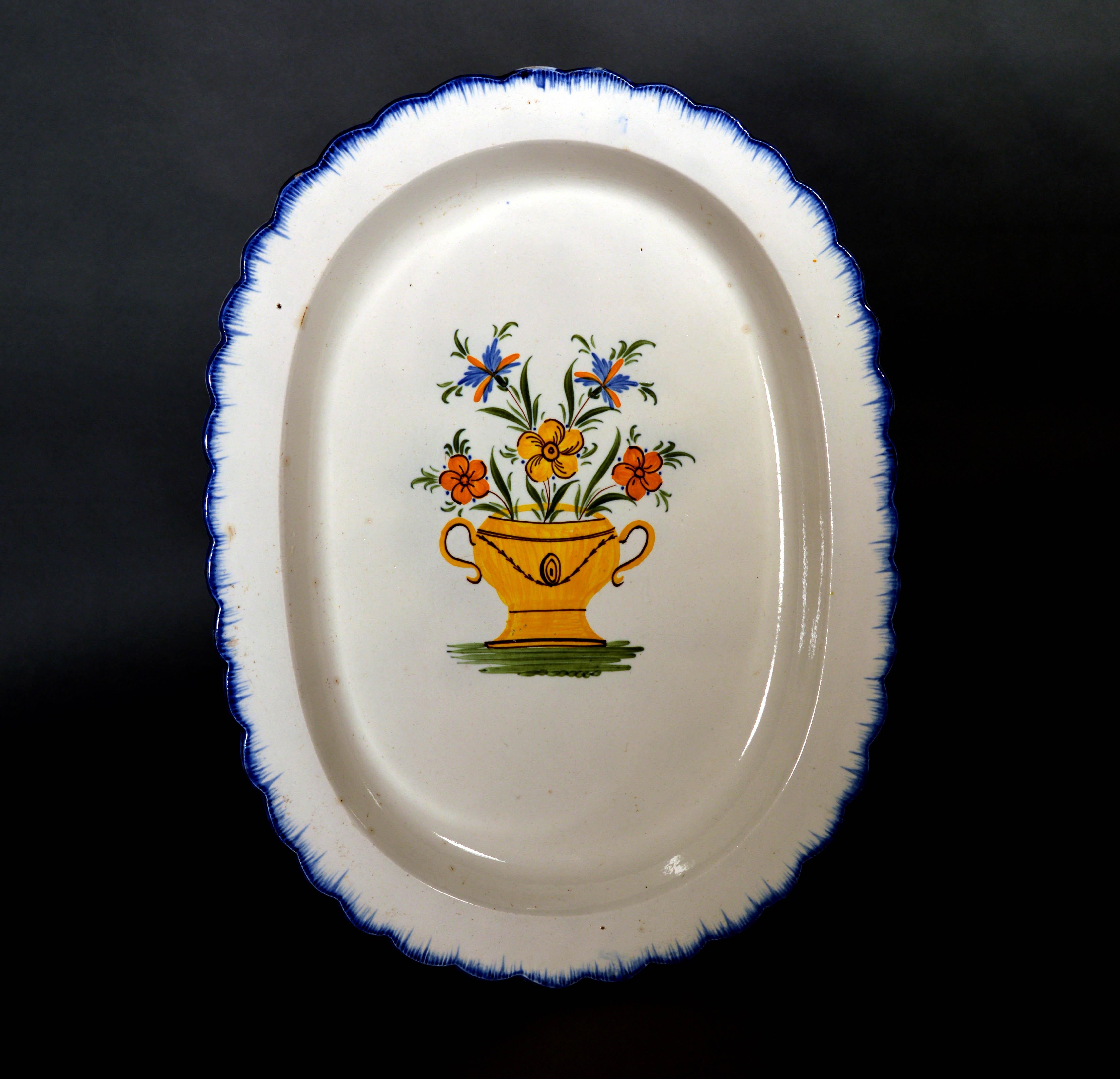 Shell-edge prattware oval dish painted with an urn of flowers,
1800-1820

The oval deep dish has a shaped border in blue with a painted blue shell-edge design. The central well is painted with a large yellow urn with brown details at the border foot