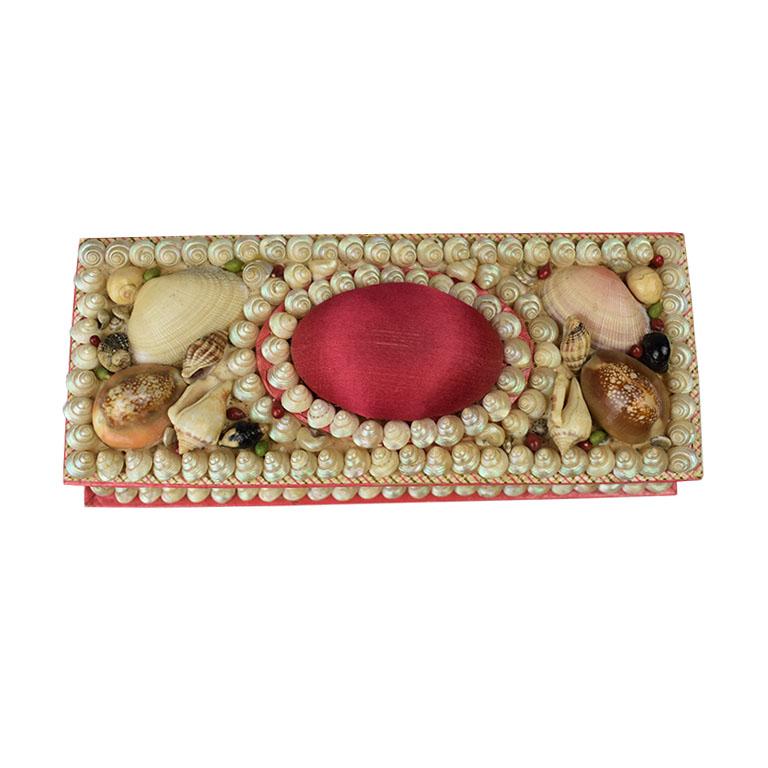 Long rectangular shell encrusted box. Carefully applied shells decorate the top and bottom edges of this red board box in a variety of colors. At the top, a cushioned upholstered red silk cushion takes center stage. At sides, are faux tiles in a