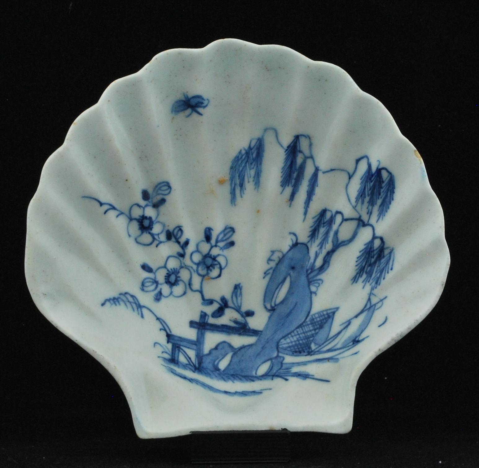 Shell dish, circa 1746-1748: Of scallop shell form on two peg feet; the interior painted in blue underglaze with a willow, rockwork, fence and flowering branches, a boat to the right, and an insect above; all a free adaptation from the Chinese