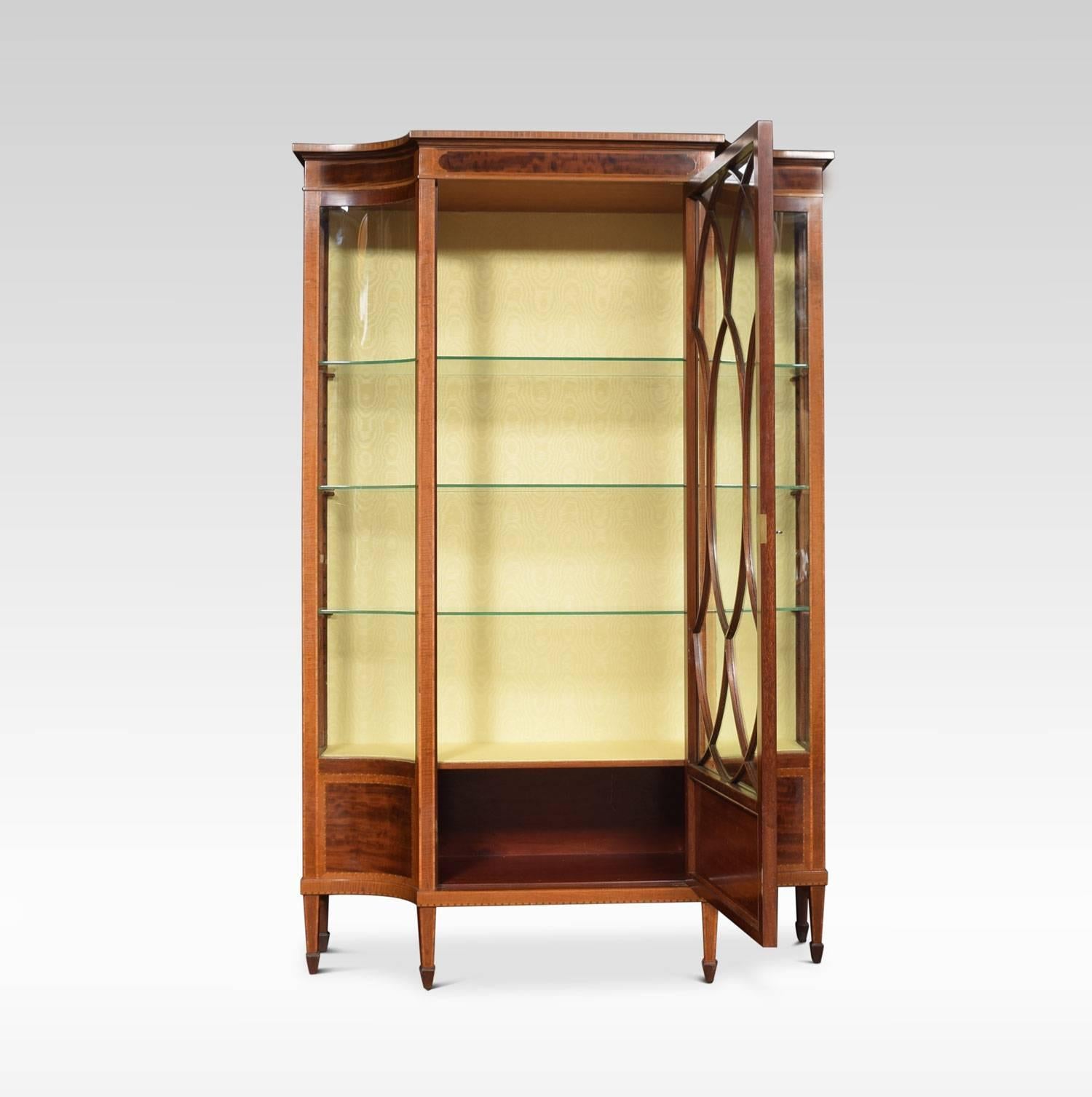 Late 19th century mahogany Sheraton revival display cabinet the shaped top above large central door opening to reveal three glazed shelves flanked by concave glazed panels above conforming undertier, all raised up on square tapering legs and spade